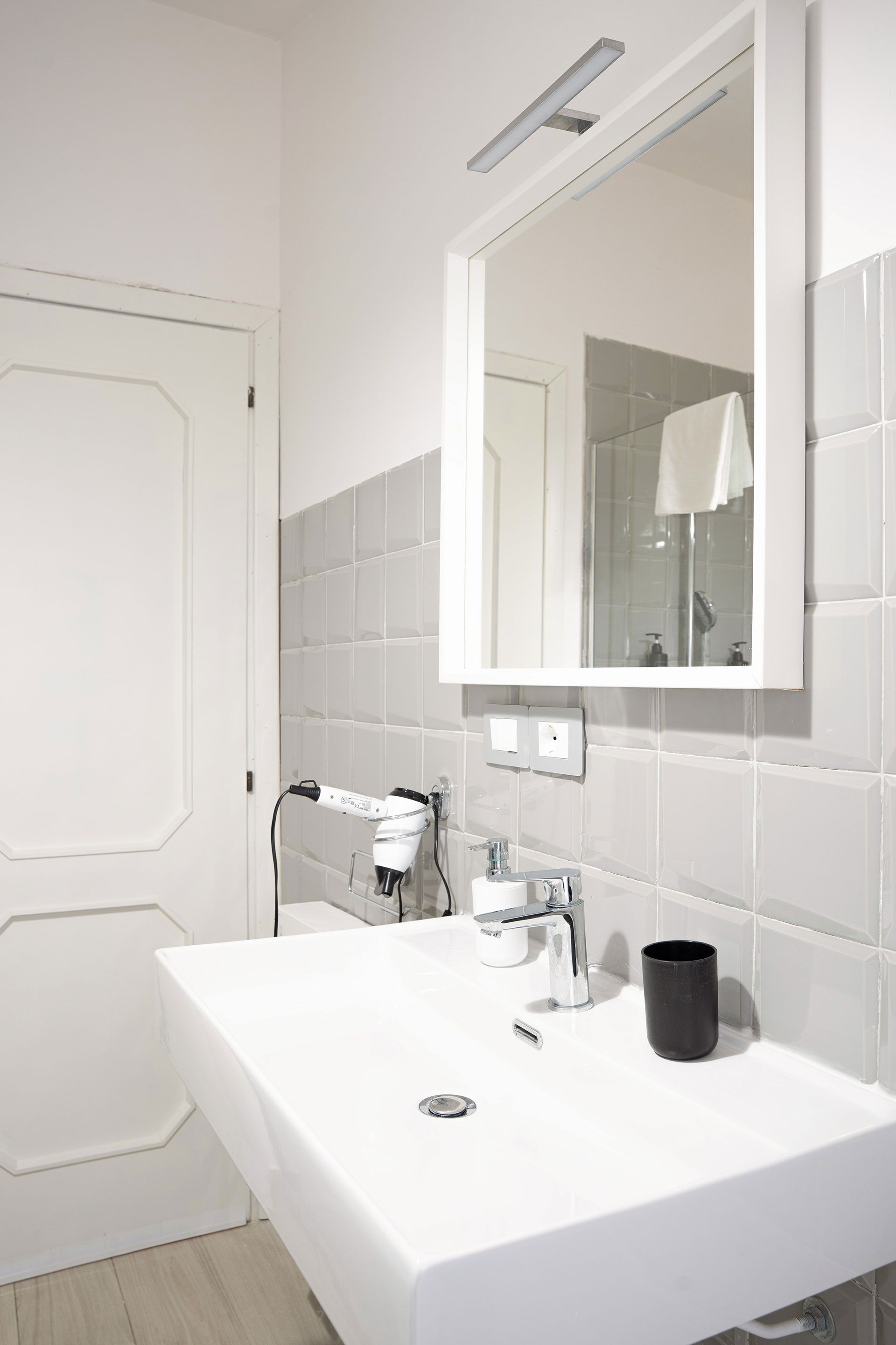 Mobile bagno luminoso con lavabo bianco, specchio, piastrelle grigie e una tazza nera sul ripiano.