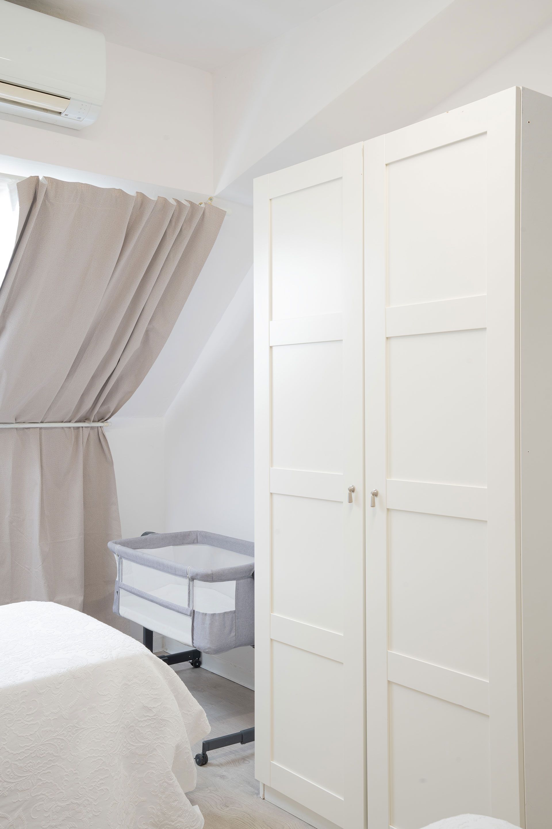 Armadio bianco in una camera da letto luminosa, accanto a una finestra con tende e un tavolino.