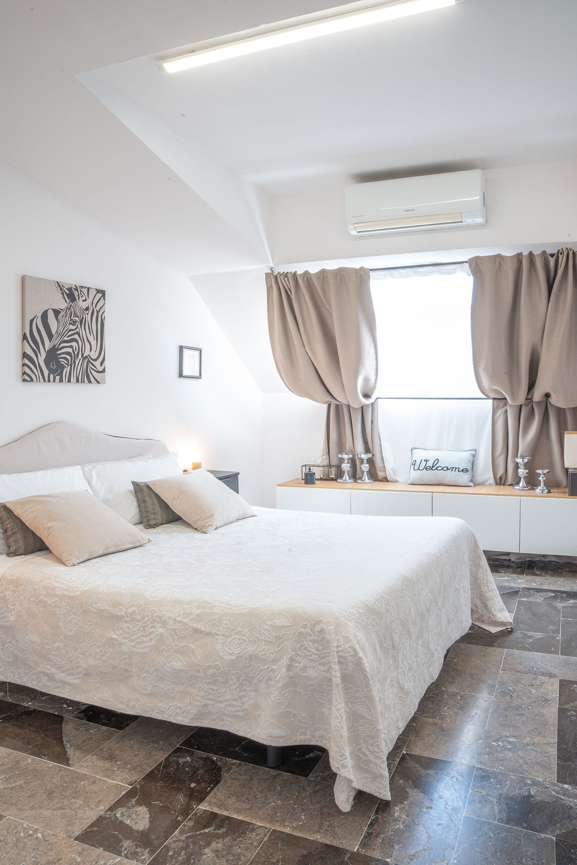 Camera da letto luminosa con letto bianco, tende beige e condizionatore d'aria sopra la finestra.
