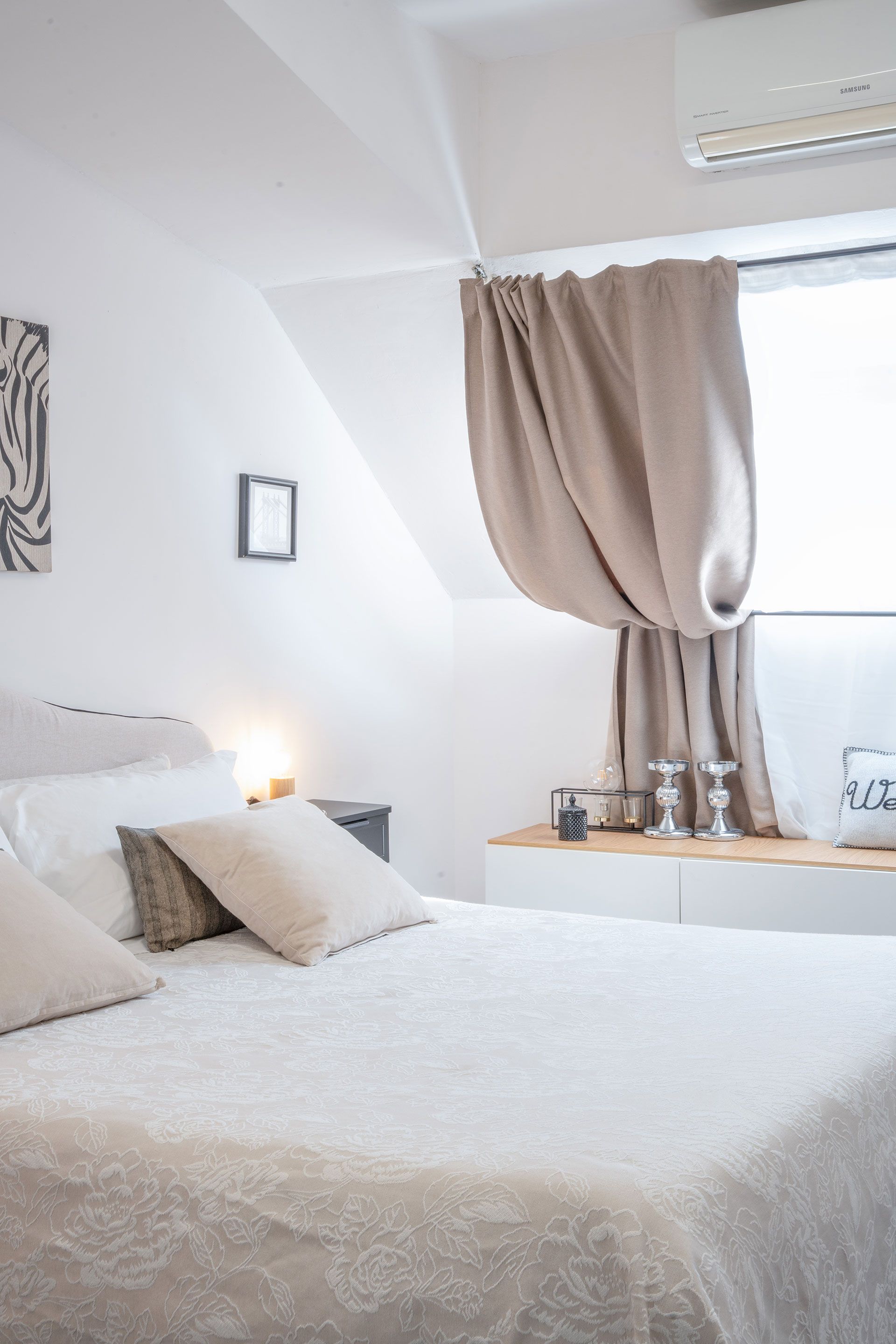 Camera da letto minimalista bianca con letto ben rifatto, cuscini beige e finestra con tenda.