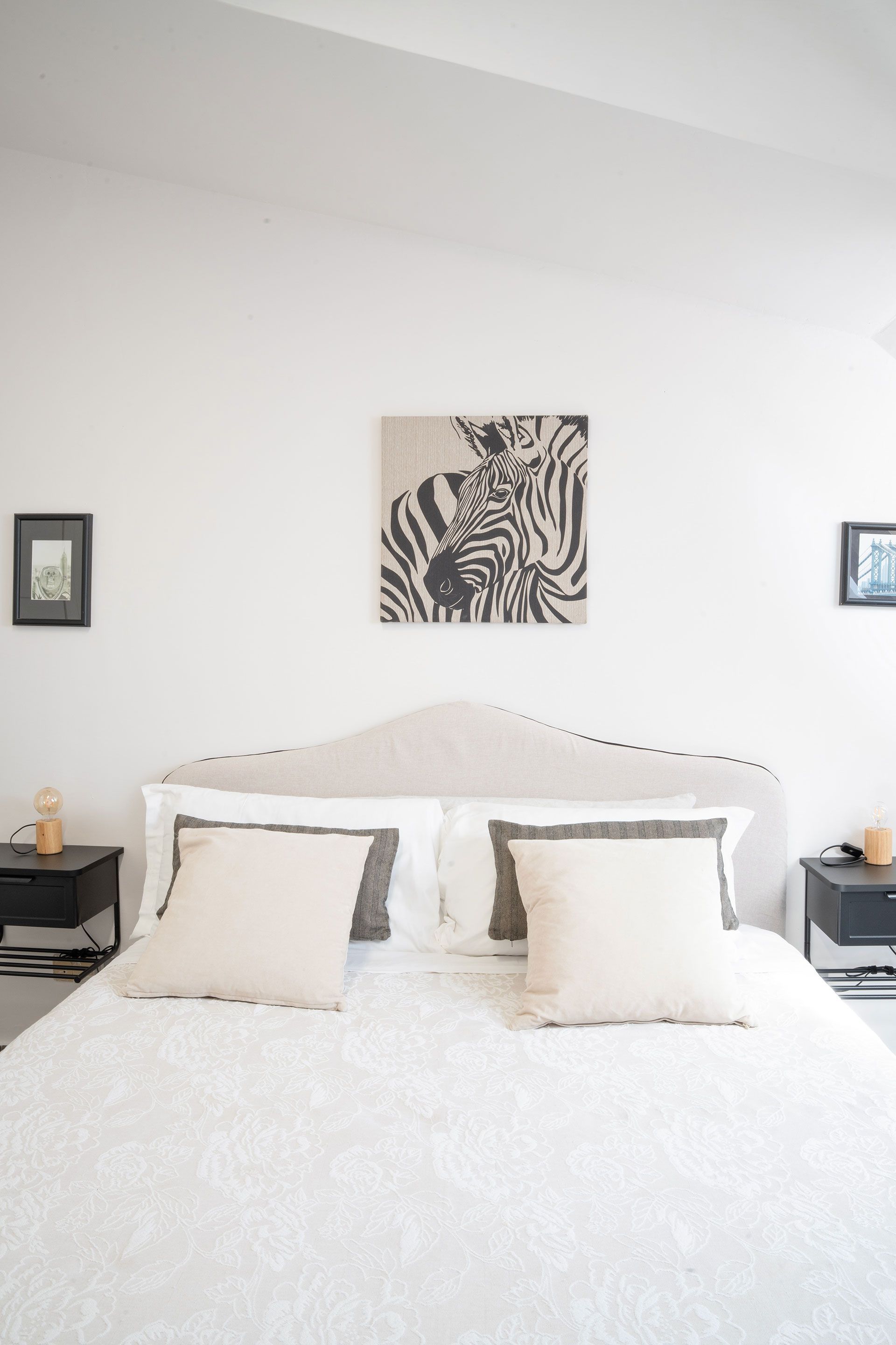 Camera da letto luminosa e bianca con letto ben rifatto, cuscini beige e quadro incorniciato sopra la testiera.