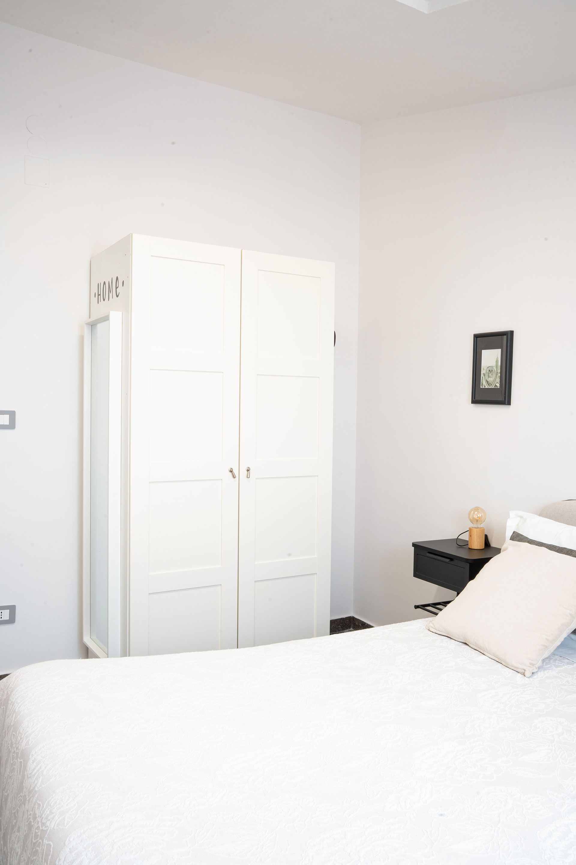 Camera da letto luminosa e bianca con letto bianco, armadio e piccolo comodino nero su una parete chiara.