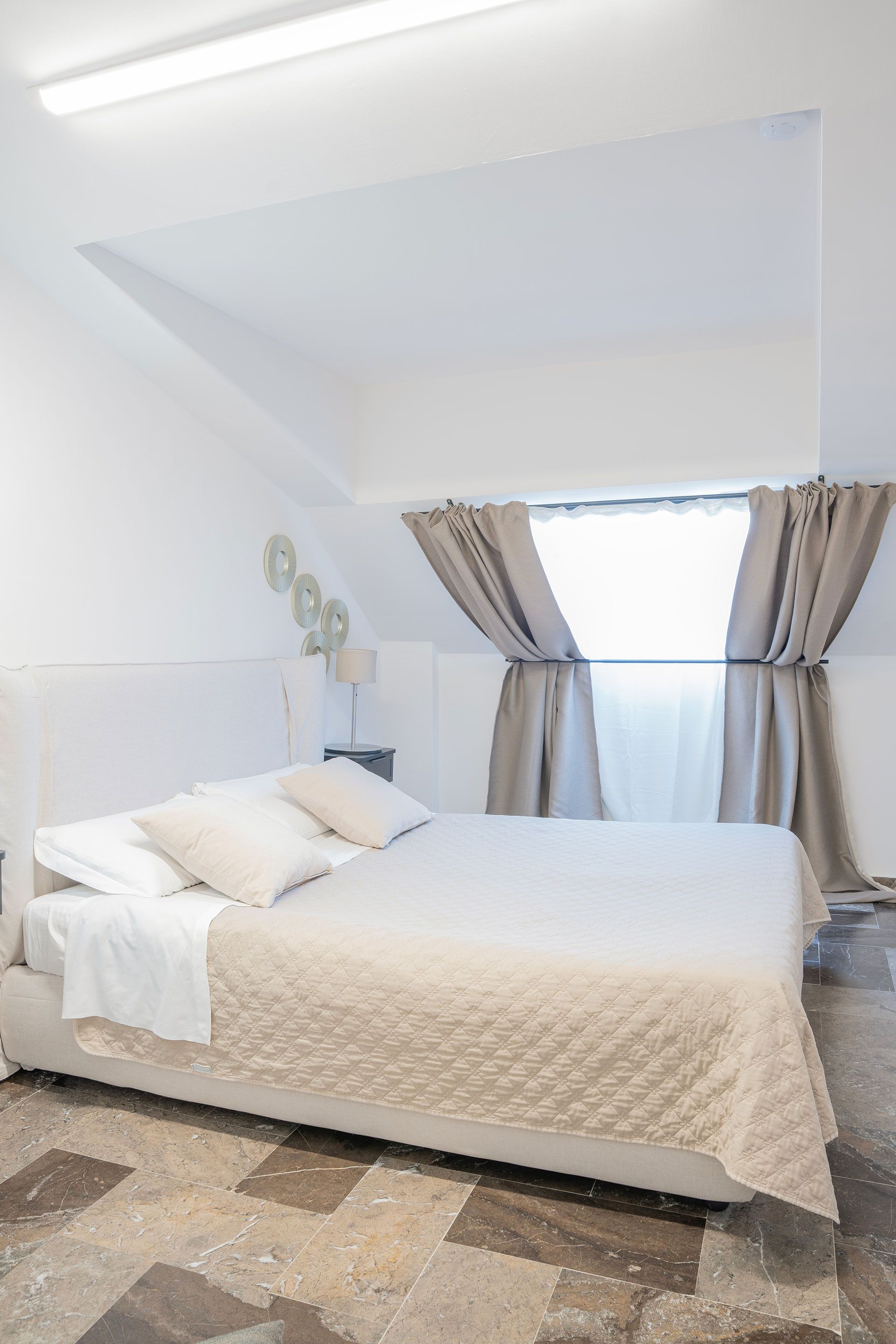Camera da letto luminosa con letto, lenzuola bianche, soffitto inclinato e finestra con tende.
