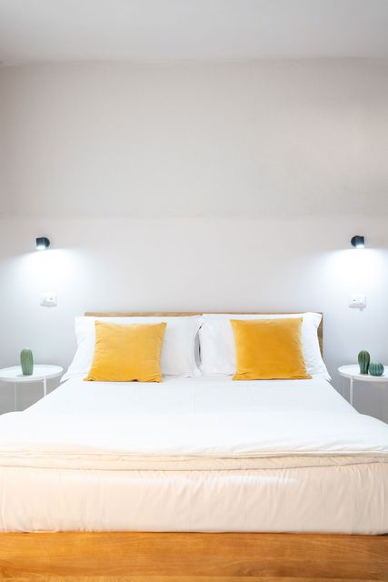 Camera da letto minimalista bianca con letto, cuscini gialli e lampade a parete.