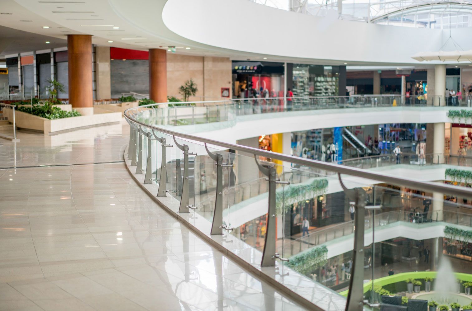 Interno moderno di un centro commerciale con ringhiera in vetro curvato, pavimenti lucidi e vetrine al piano inferiore.