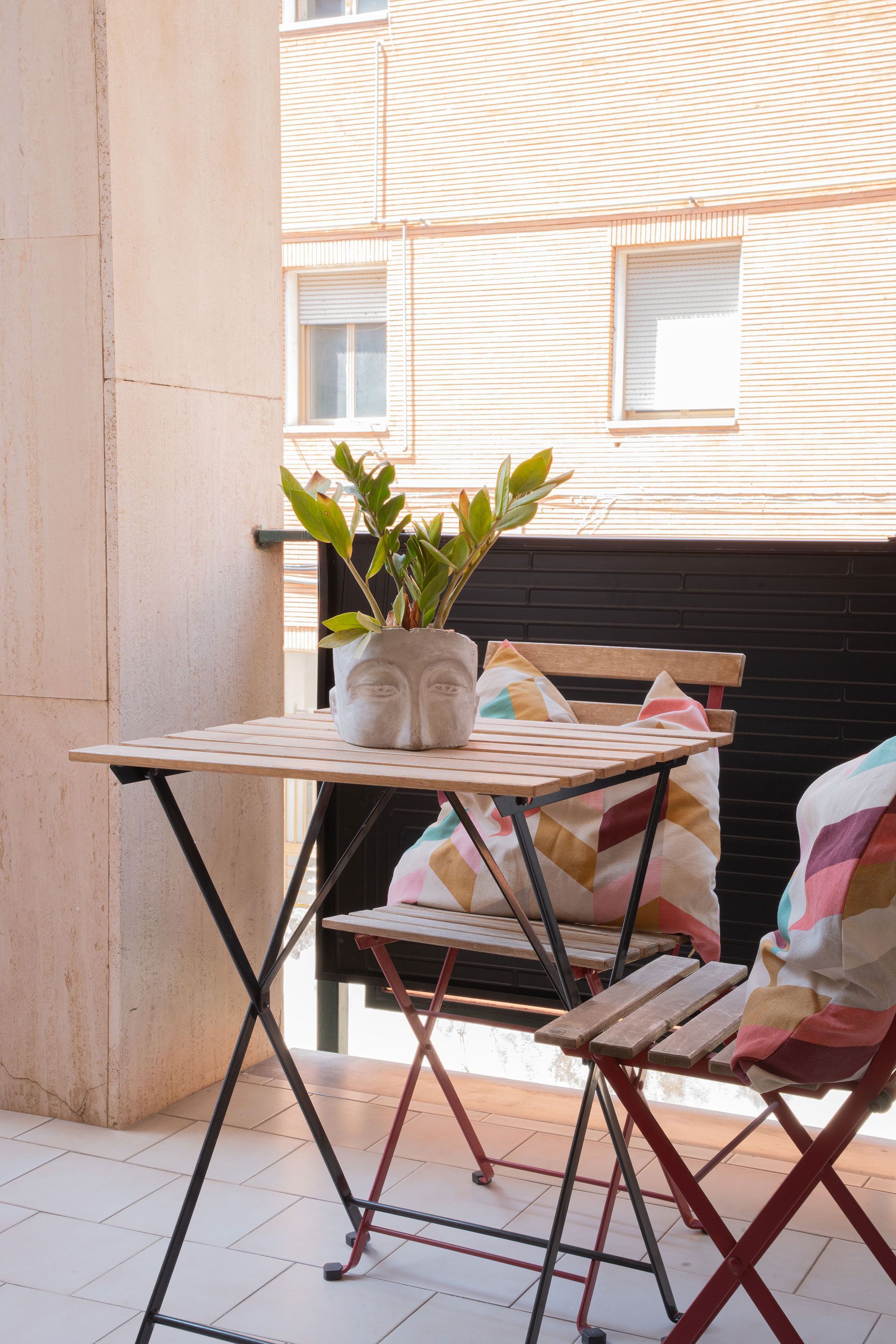 Piccolo patio con tavolo rotondo, sedie pieghevoli e un vaso di fiori accanto al muro di un edificio.