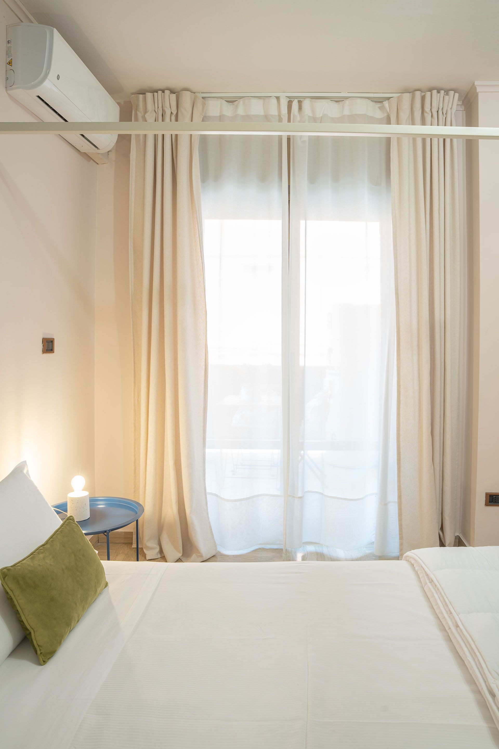 Camera da letto luminosa con lenzuola bianche, tende beige e luce del sole che filtra attraverso tende trasparenti.