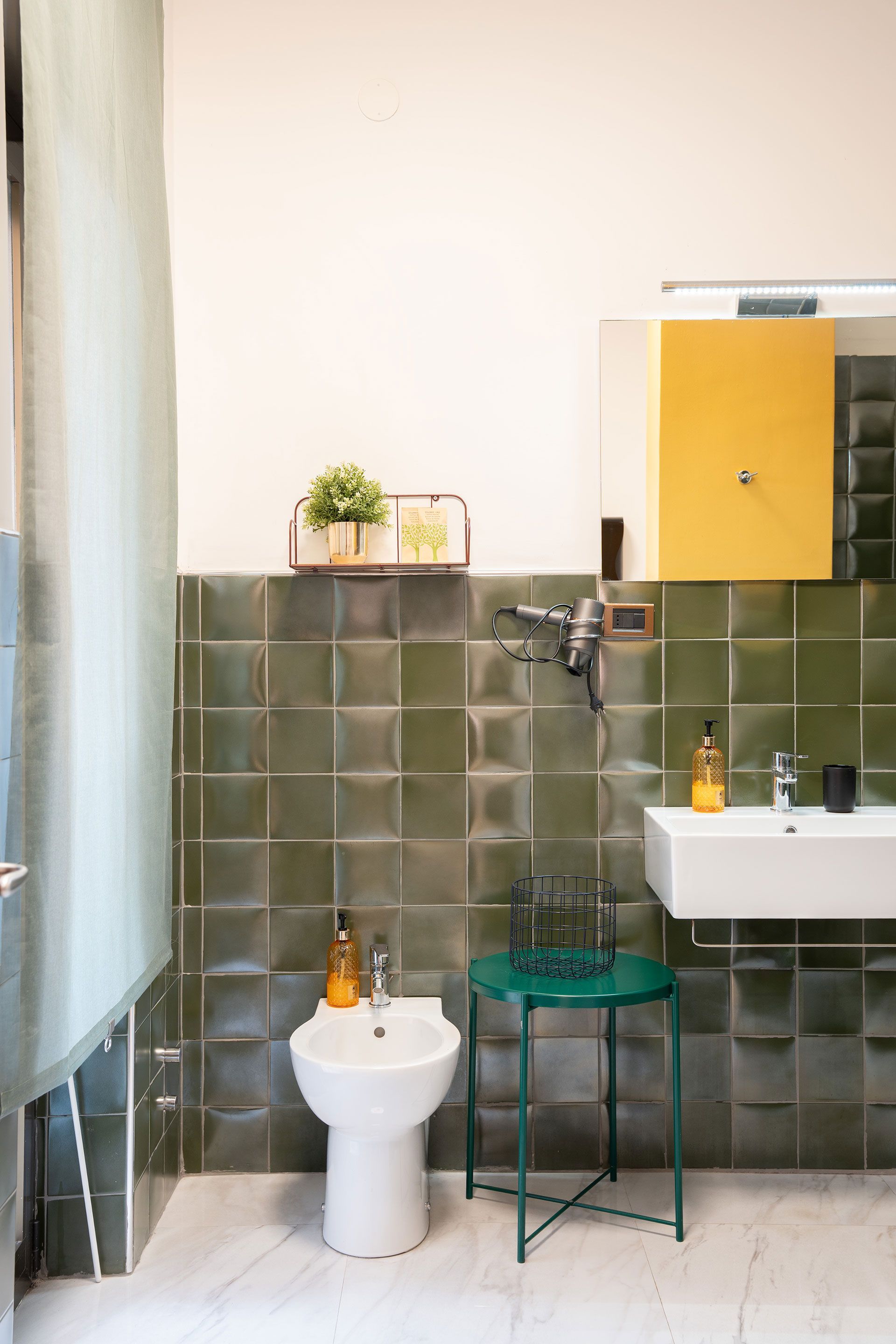 Piccolo bagno moderno con pareti rivestite di piastrelle verdi, WC bianco, lavabo, doccia e un mobiletto giallo con specchio sopra.