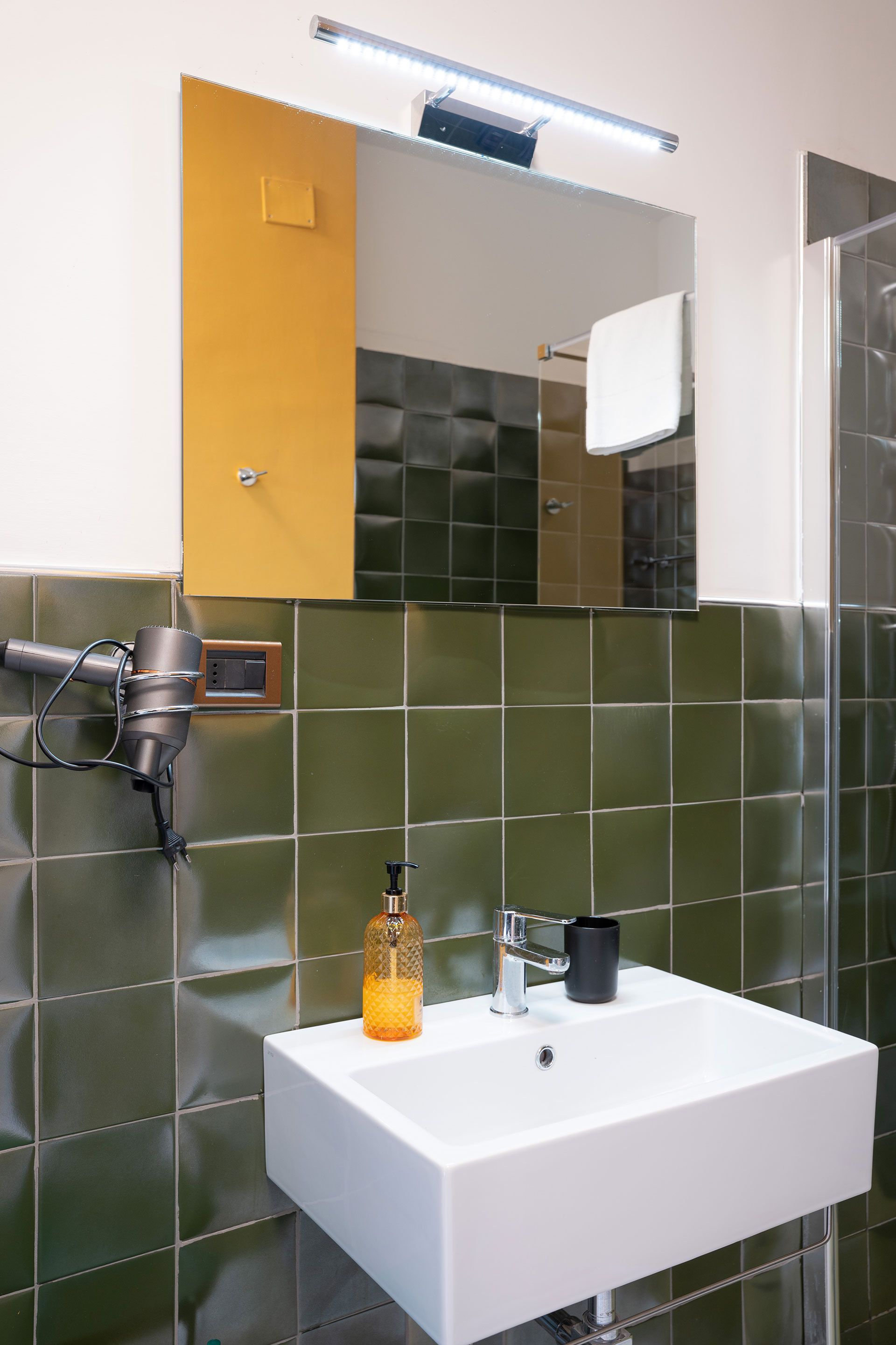 Lavabo del bagno con parete rivestita di piastrelle verdi, mobiletto con specchio, applique a parete e dispenser di sapone.