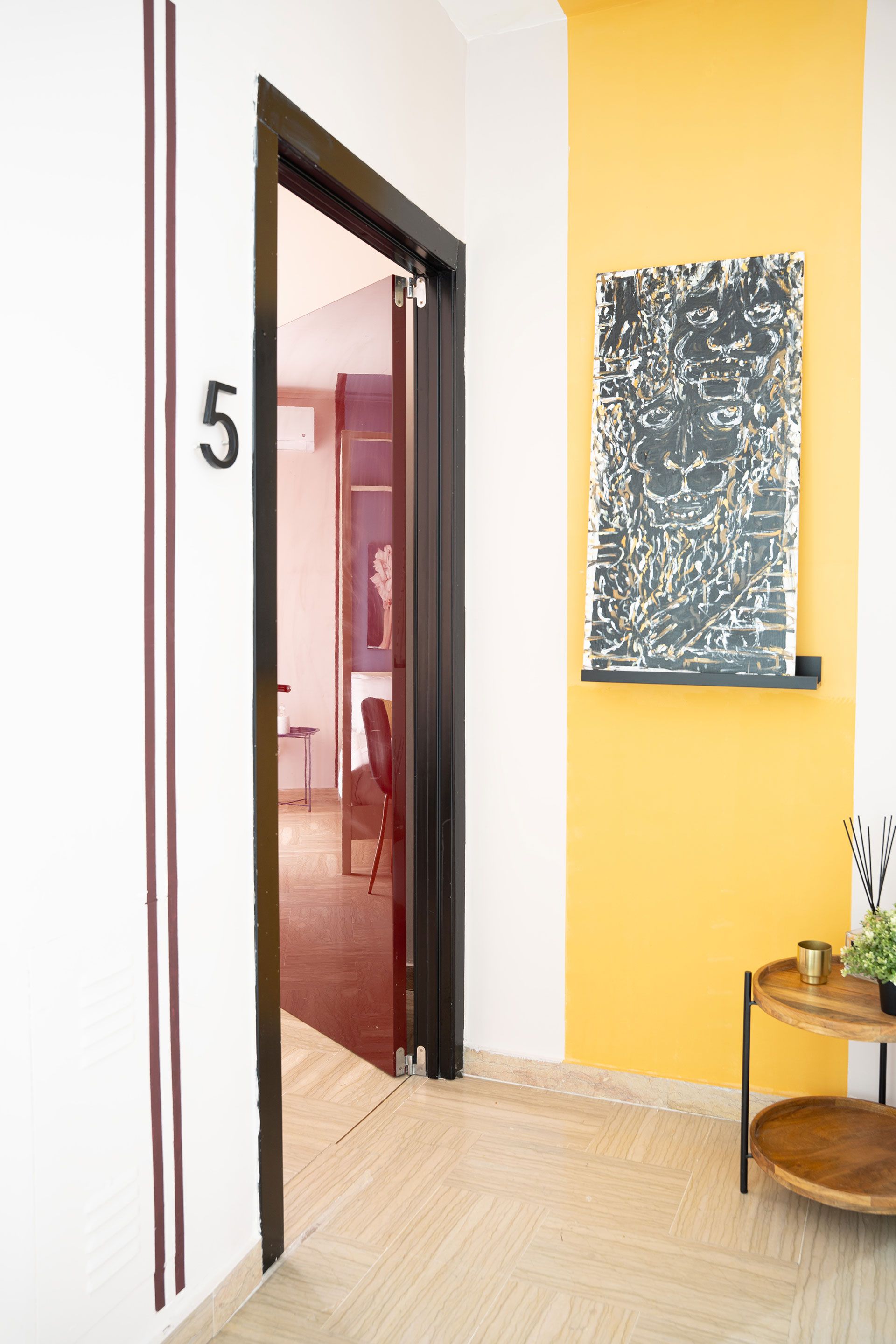 Porta numero 5 in un corridoio luminoso con pareti gialle e bianche, opere d'arte alle pareti e un tavolino.