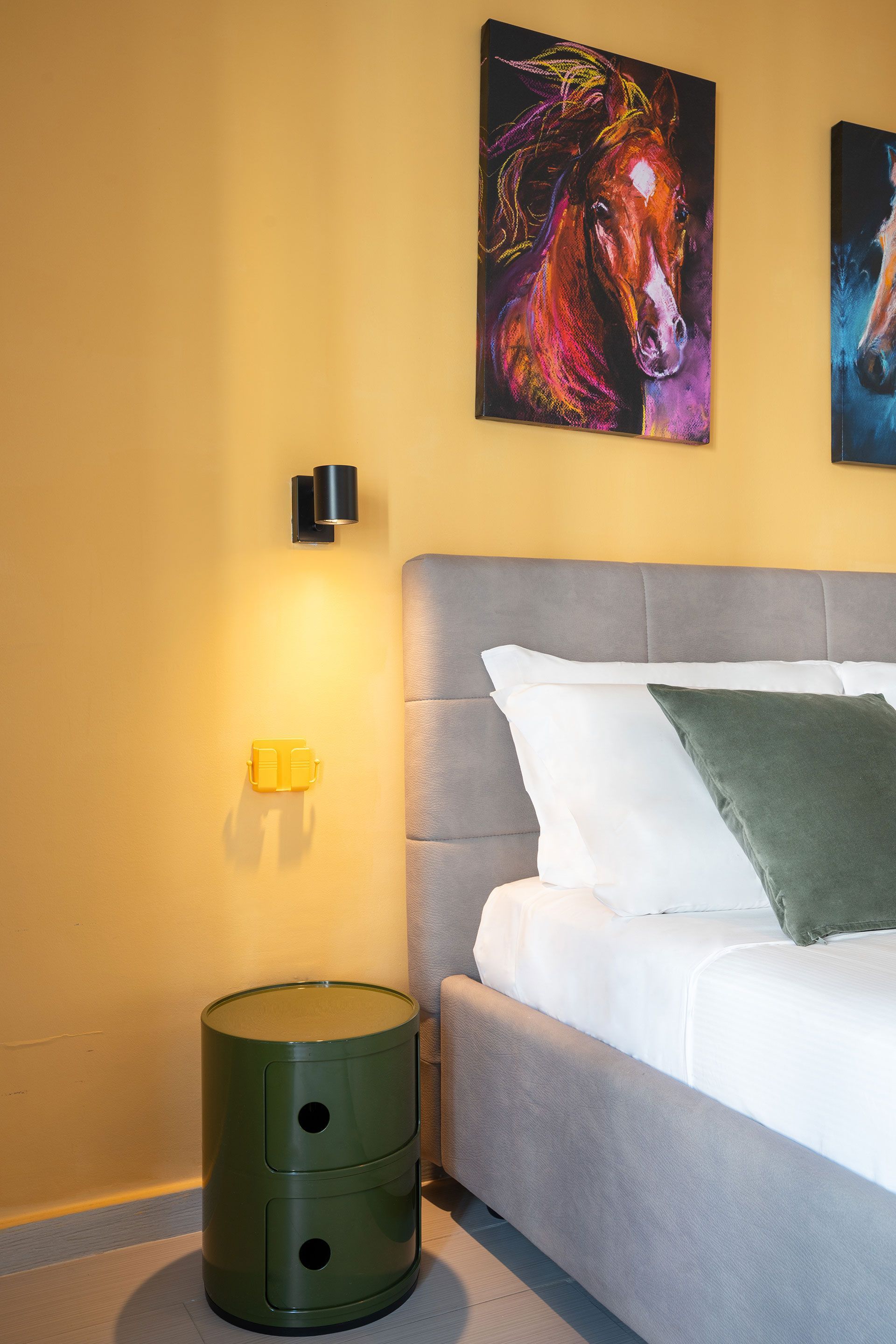 Angolo camera da letto moderno con letto grigio, parete gialla, comodino verde e quadri colorati.