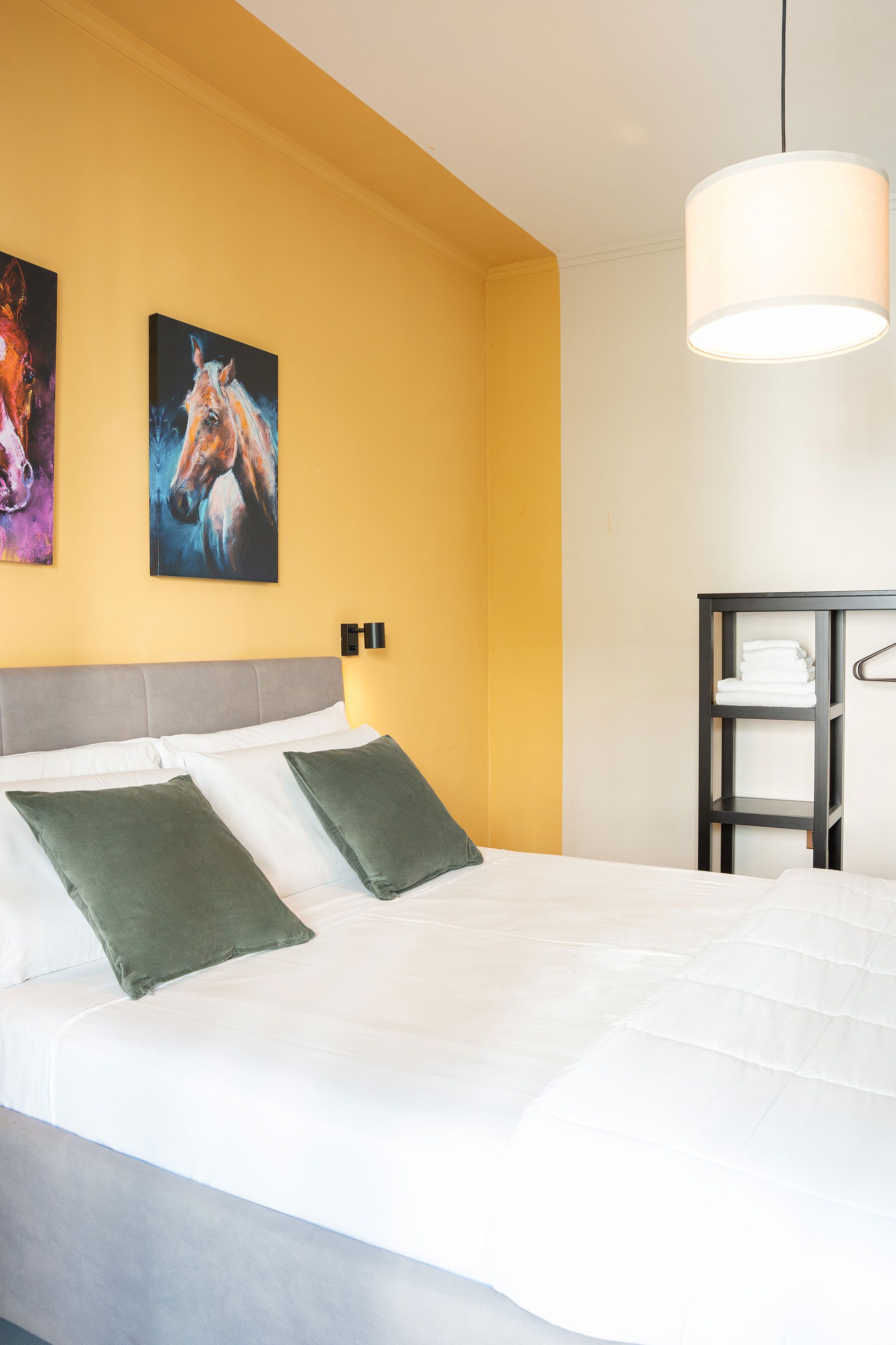 Accogliente camera da letto con letto bianco, cuscini verdi, parete decorativa gialla, quadri e lampada a sospensione.