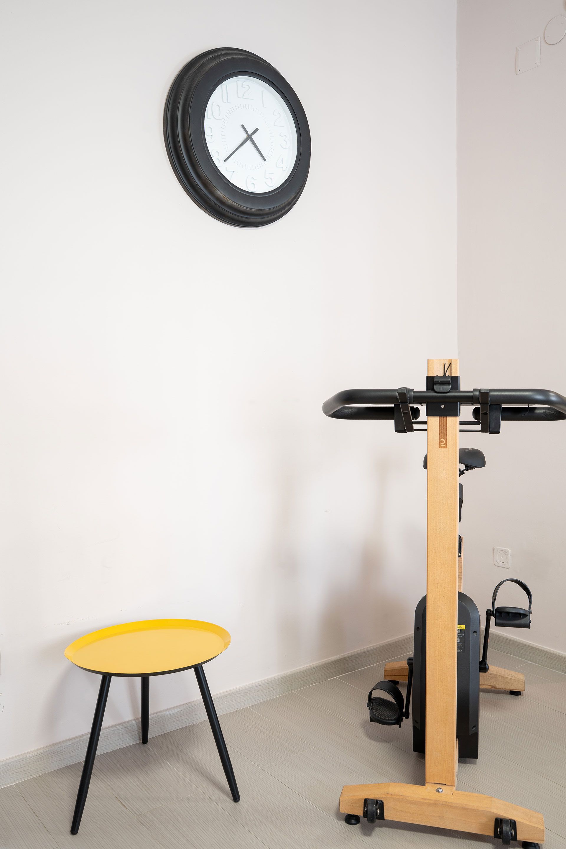 Sala fitness minimalista con orologio dalla cornice nera, sgabello giallo e scrivania in legno per lavorare in piedi.