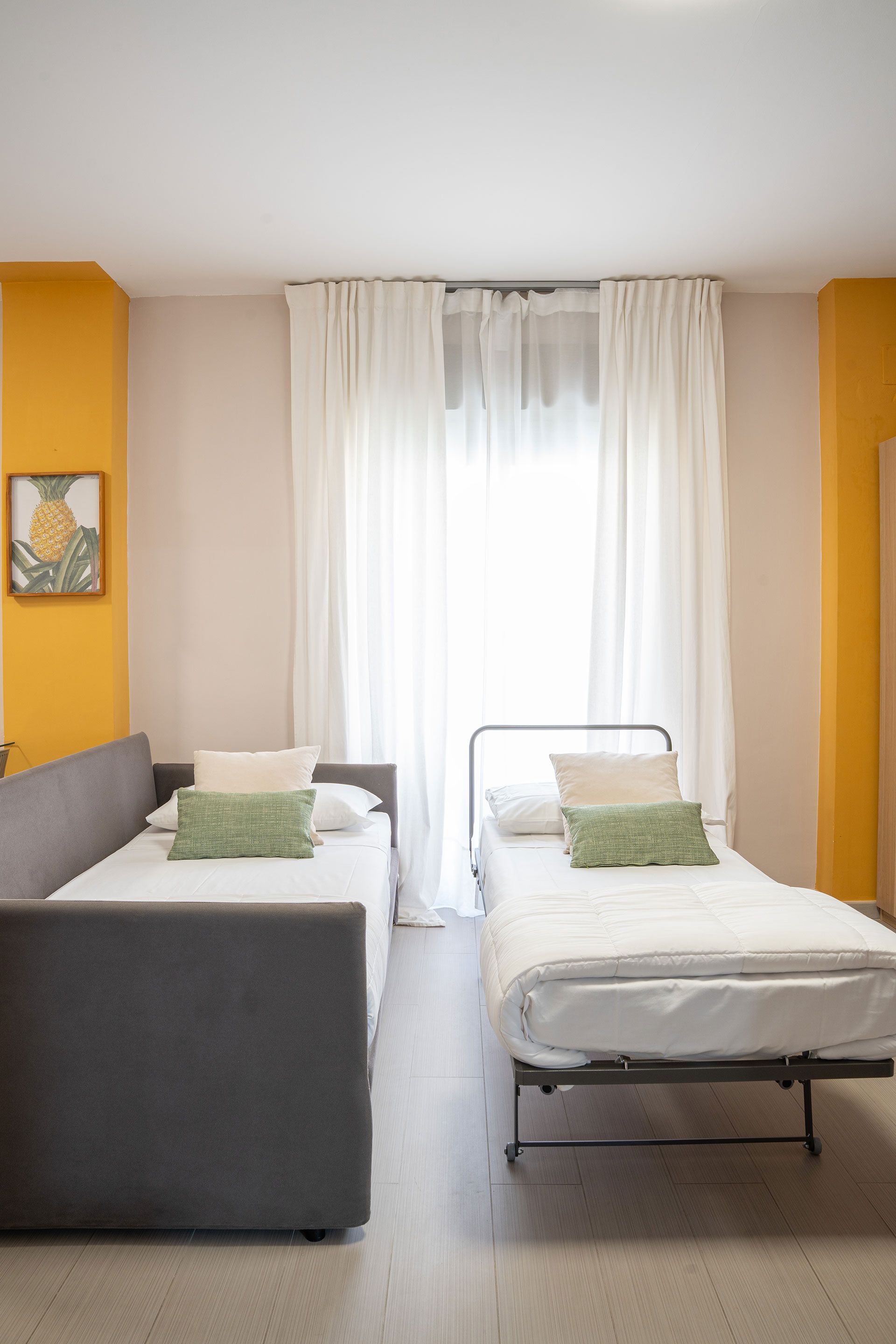 Piccola camera d'albergo con due letti, divano grigio, pareti gialle e tende trasparenti accanto a una finestra luminosa.