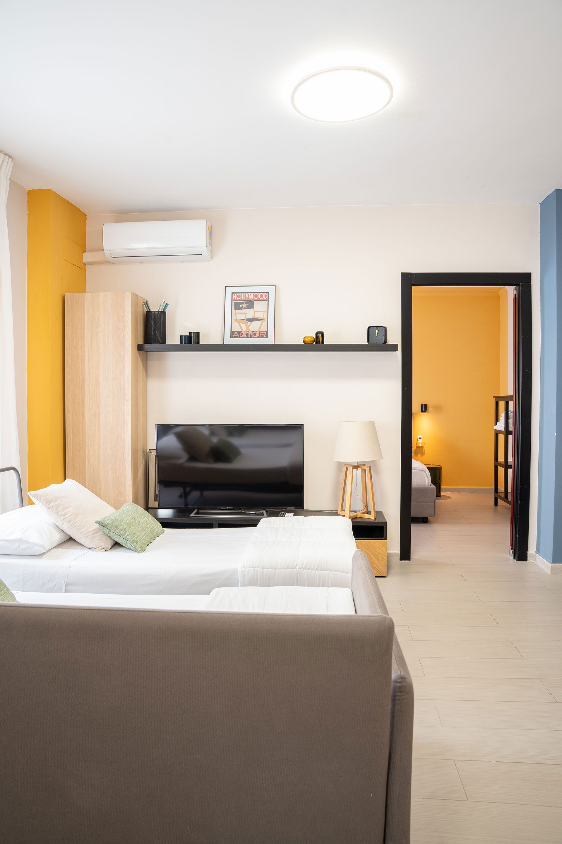 Soggiorno moderno con pareti bianche, divano grigio, TV, condizionatore e una porta che conduce a una stanza illuminata da una luce calda.