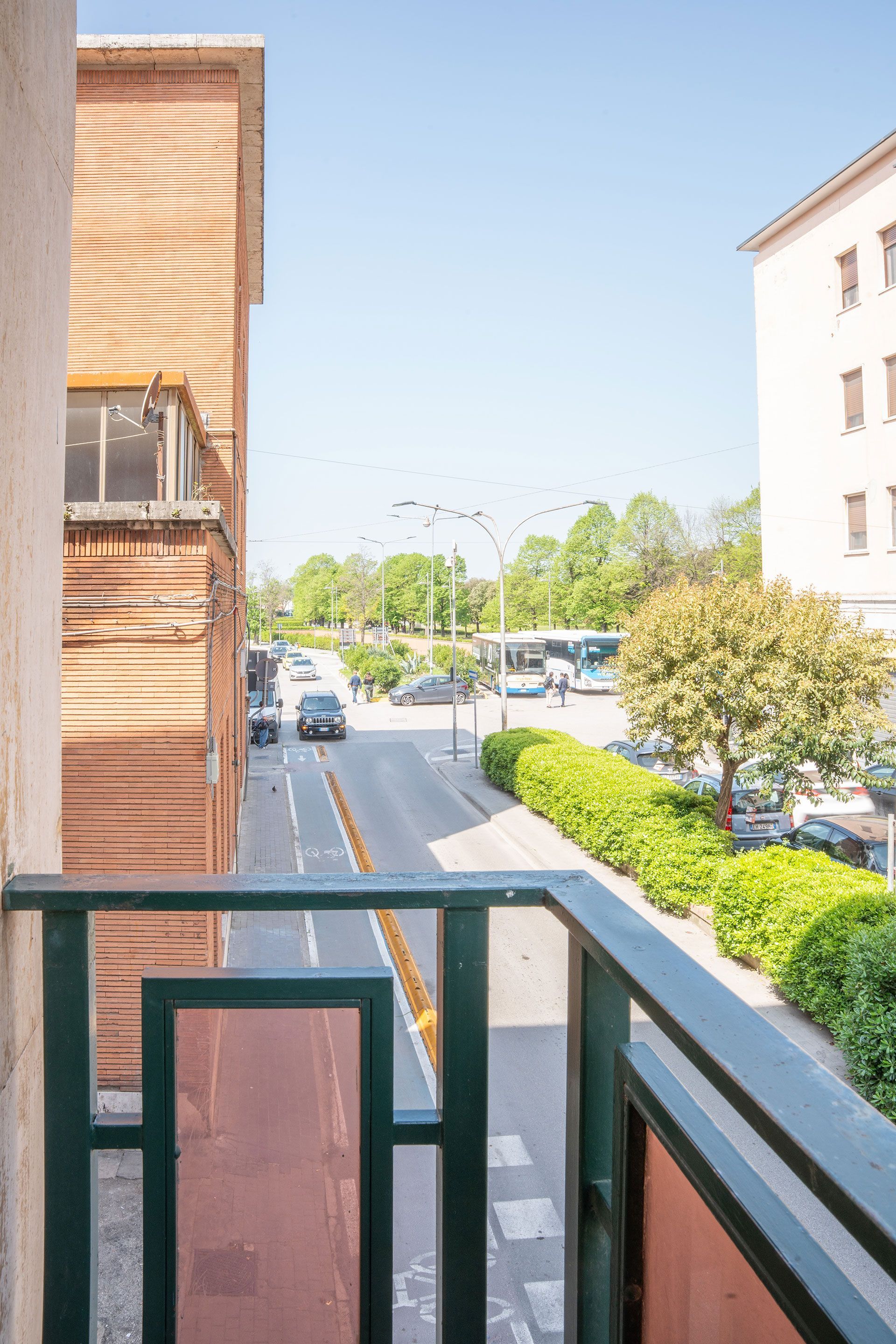 Vista dal balcone su una strada soleggiata con auto parcheggiate, alberi ed edifici su entrambi i lati.