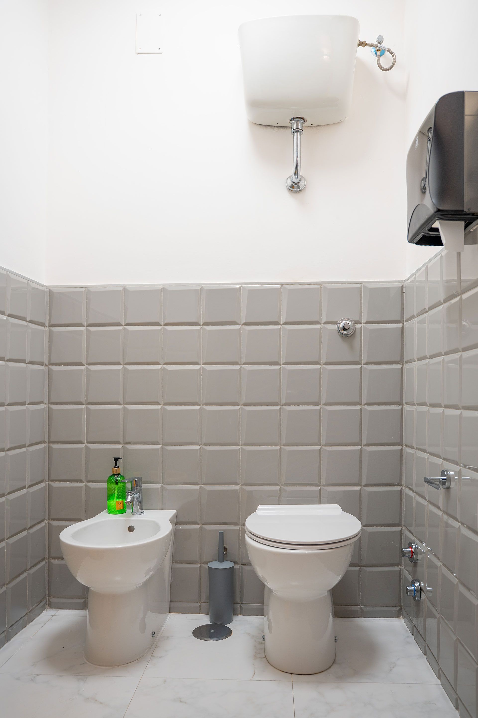 Piccolo bagno piastrellato con WC, bidet e cassetta di scarico a parete, su pareti bianche e grigie.