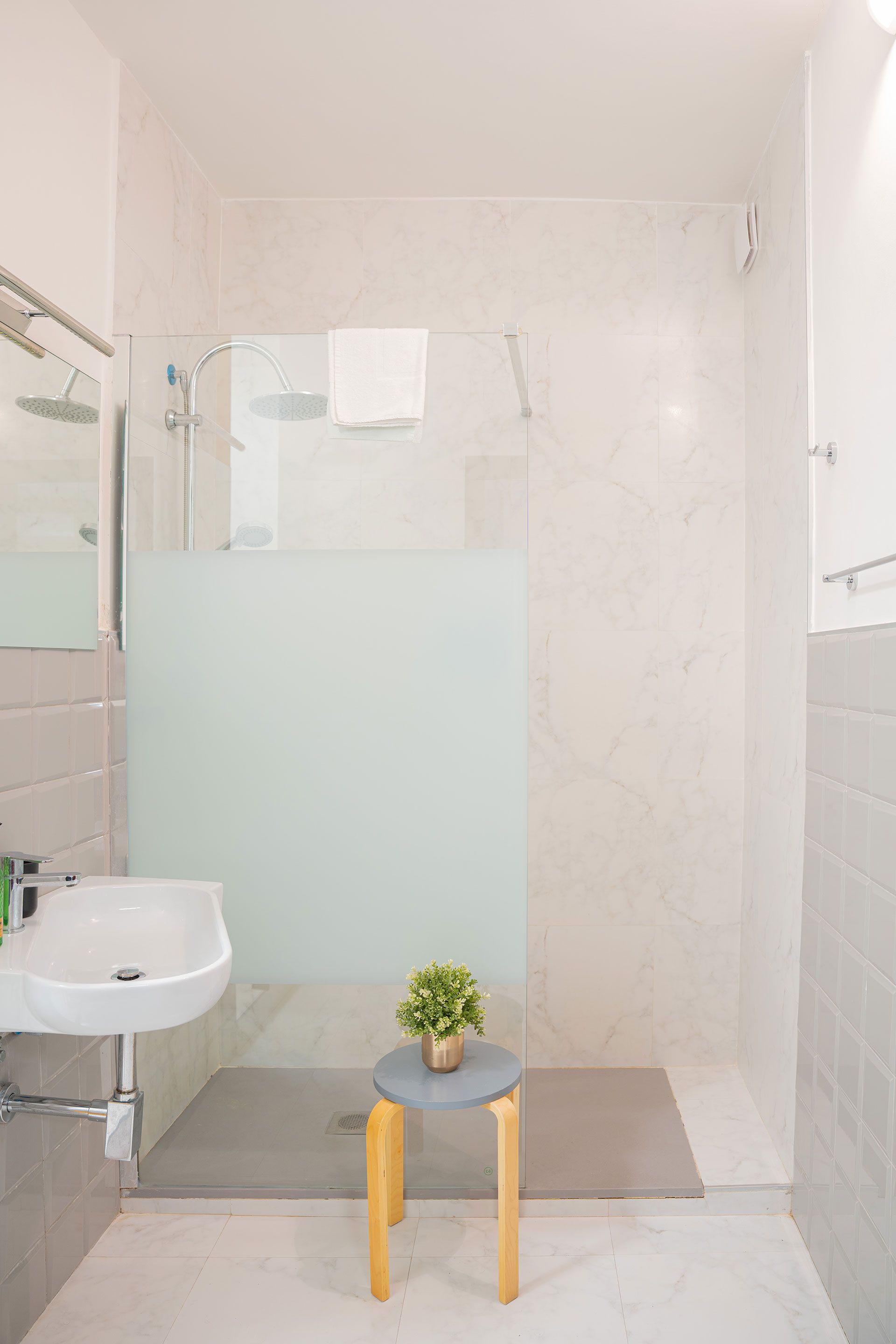 Piccolo bagno moderno con box doccia in vetro satinato, lavabo a parete e sgabello con pianta in vaso.