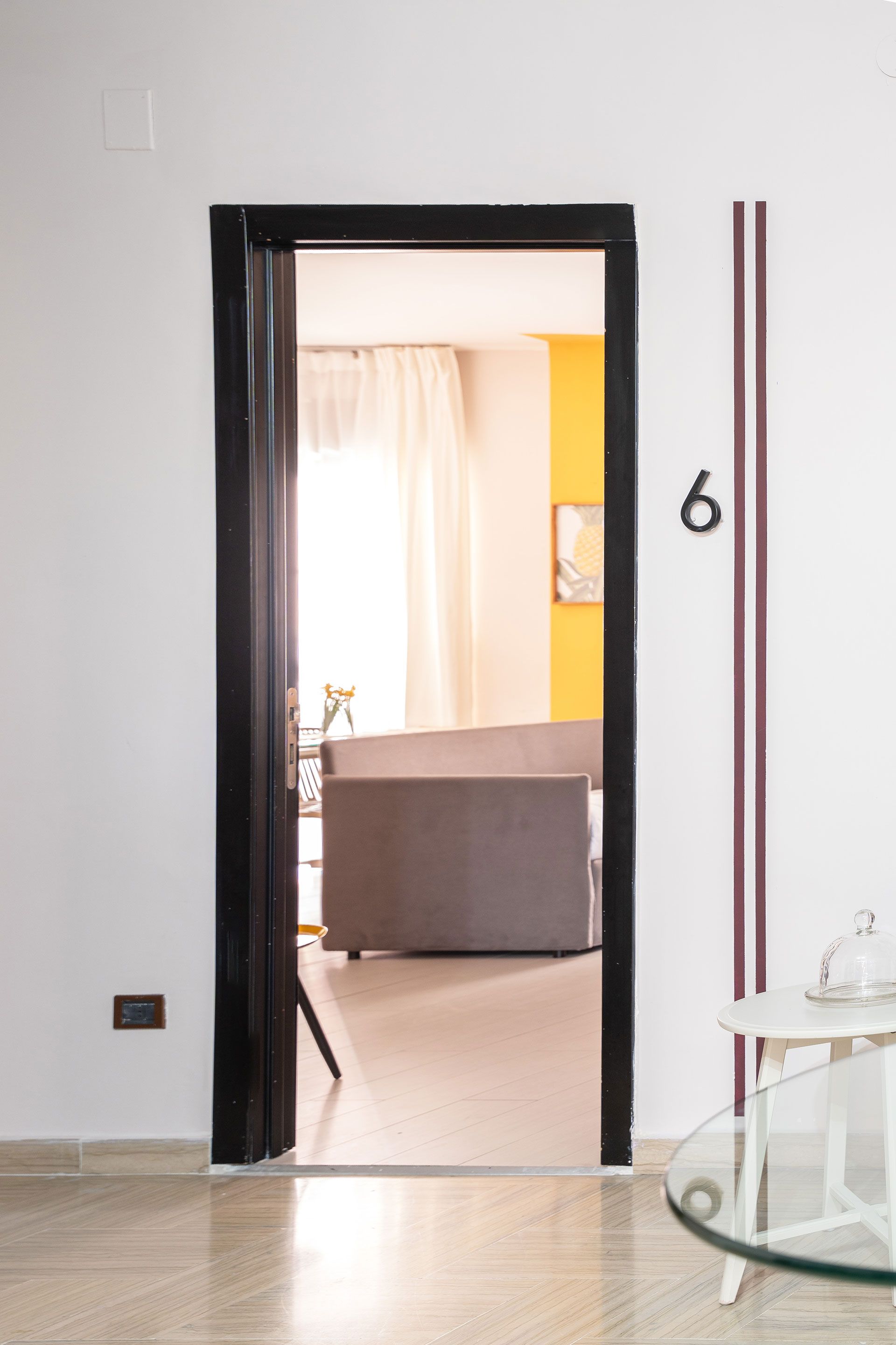 Ingresso di un soggiorno moderno con finiture nere, pareti bianche, tavolo in vetro e un divano grigio oltre la porta.