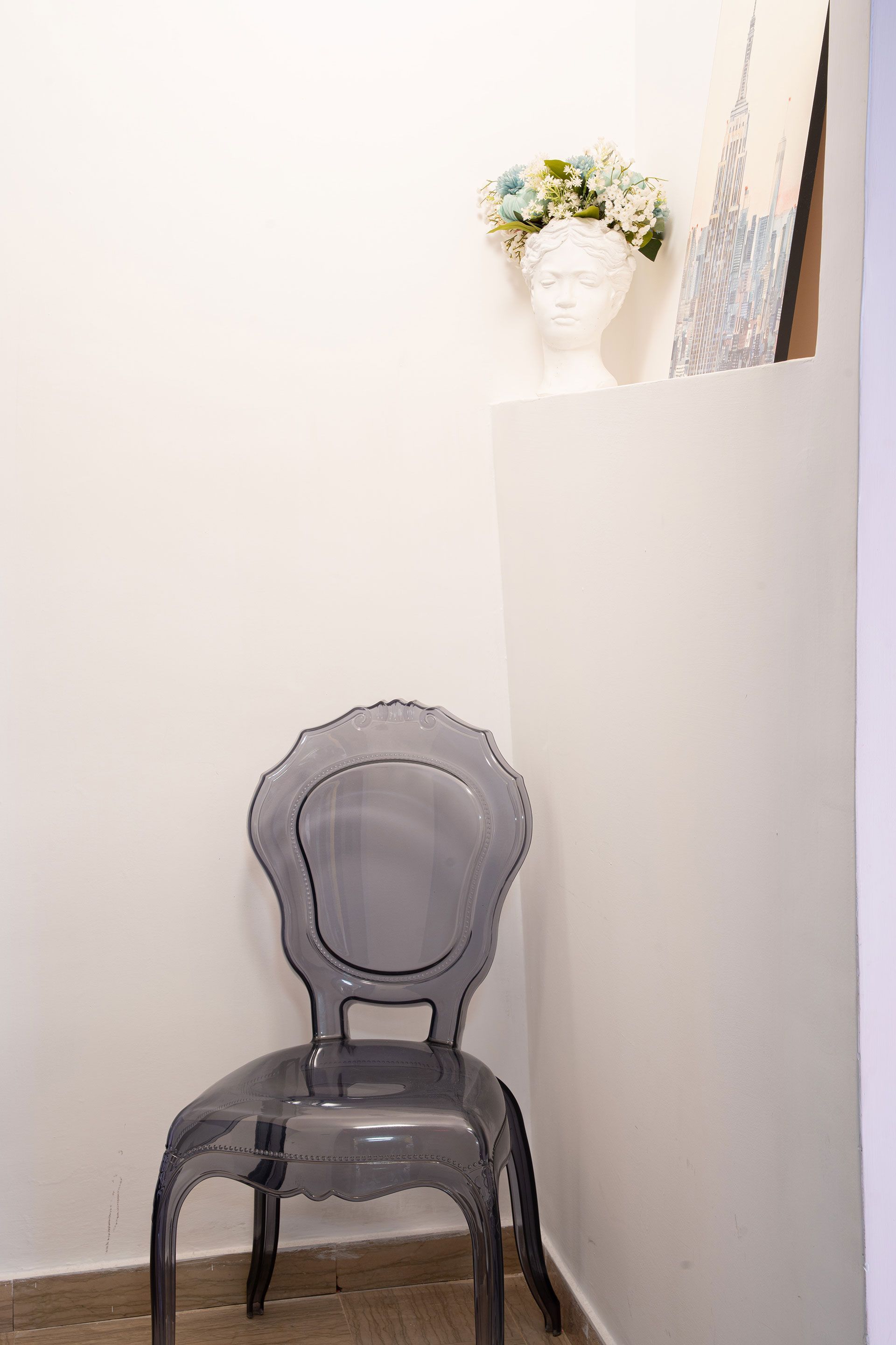 Sedia trasparente in un angolo bianco accanto a un muretto con una testa di manichino floreale e un'opera d'arte sopra.