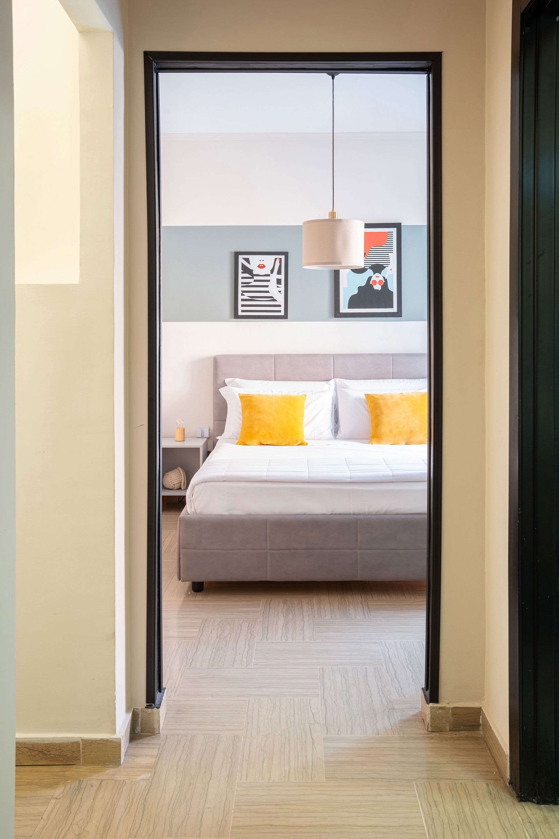 Uno scorcio attraverso la porta di una camera da letto moderna con un letto grigio, cuscini gialli e quadri appesi alla parete.
