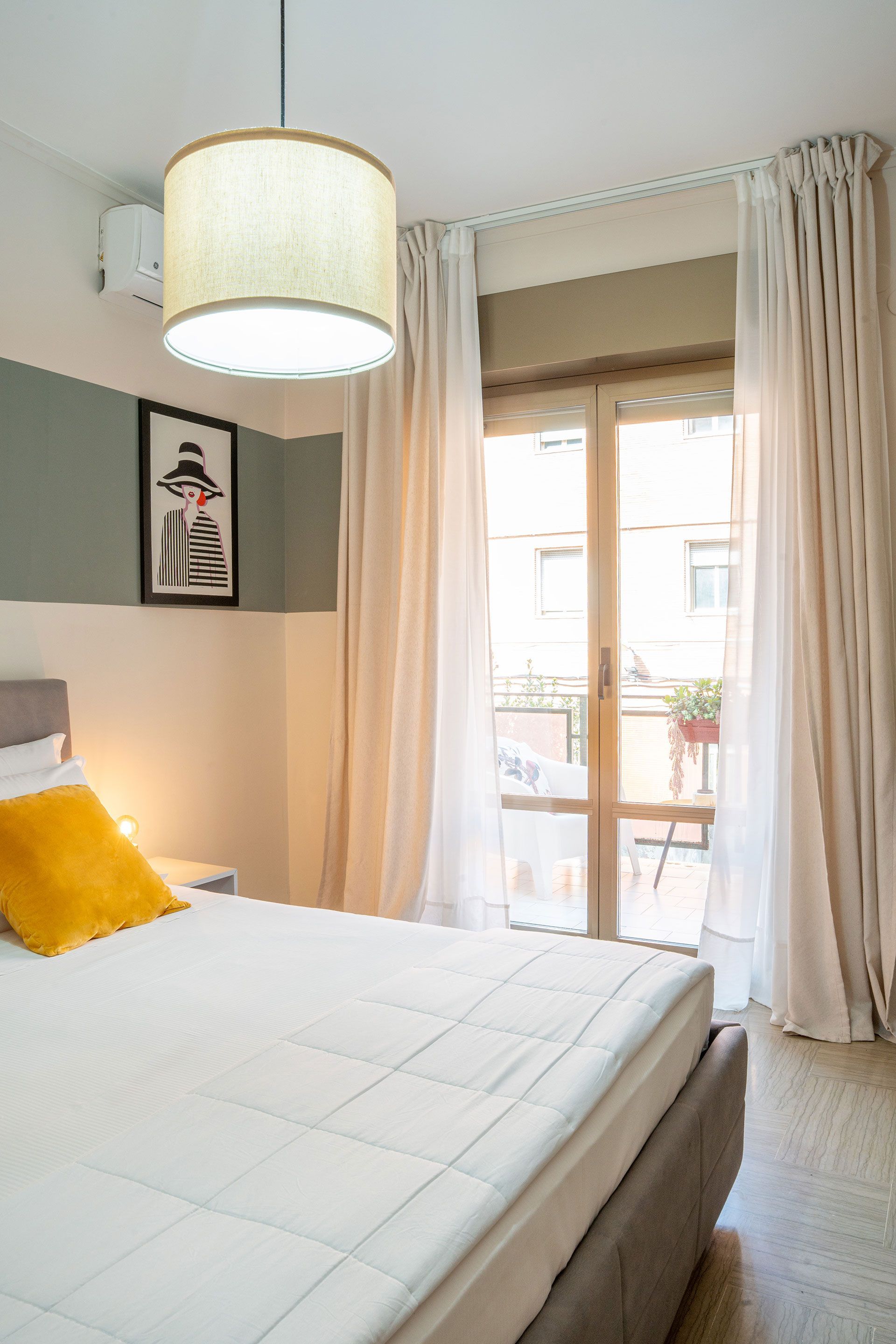 Camera da letto luminosa con letto bianco, cuscino giallo, tende color crema e lampada a sospensione accanto alla porta del balcone.