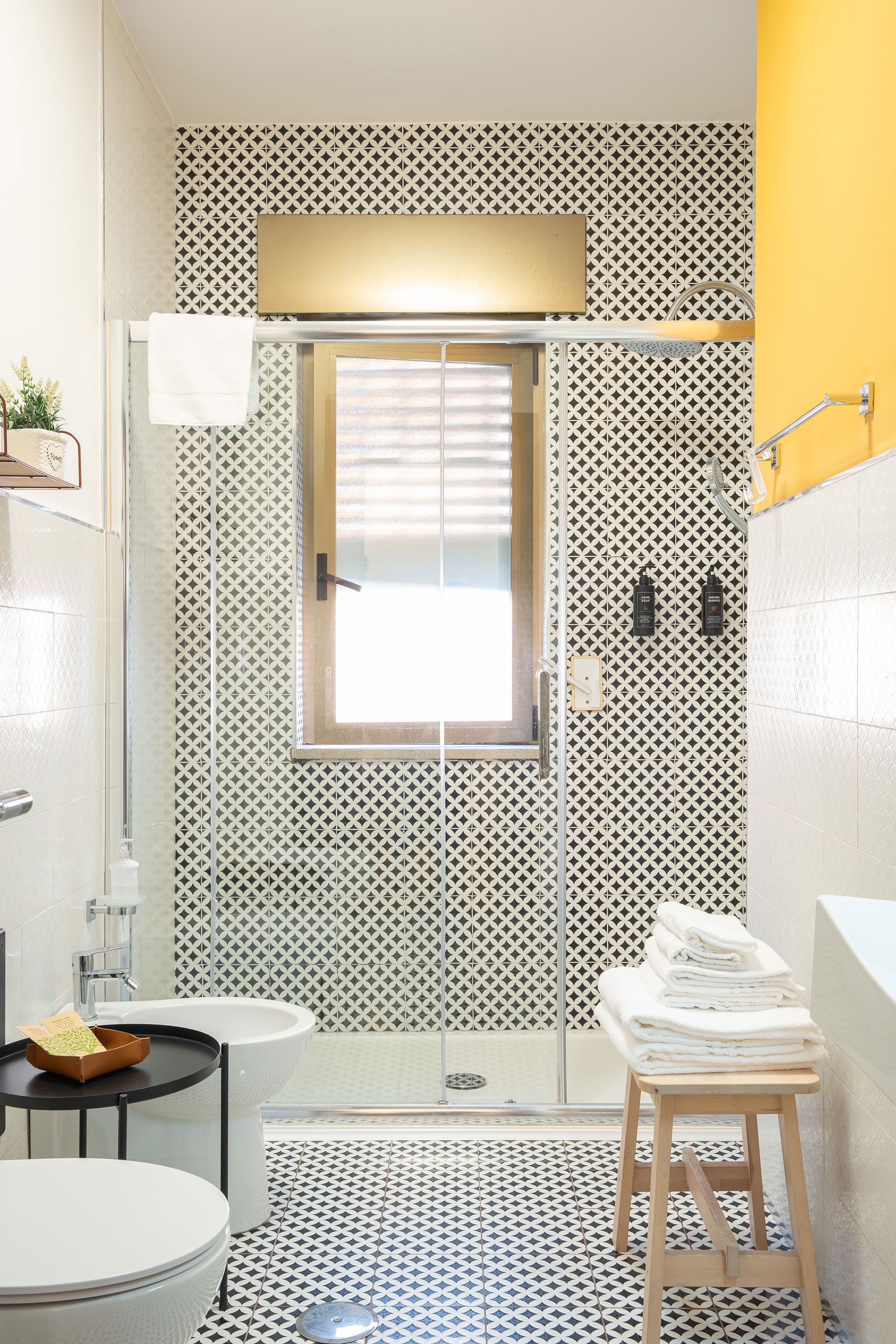 Luminoso bagno rivestito con piastrelle a mosaico, dotato di doccia, finestra, specchio a parete e asciugamani bianchi su uno sgabello.
