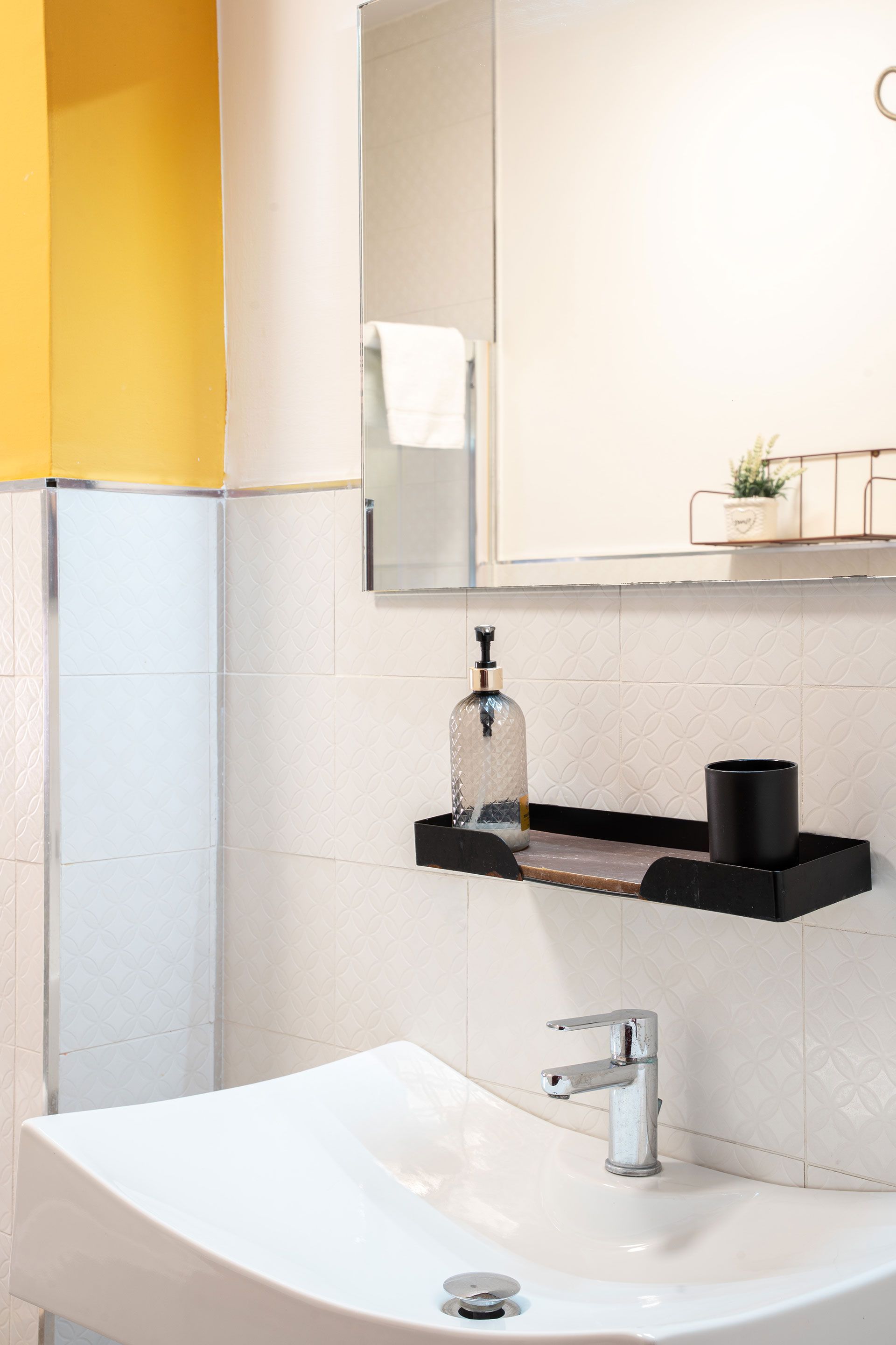 Lavabo da bagno di piccole dimensioni con specchio, mensola nera, dispenser di sapone e piastrelle a parete di colore chiaro.