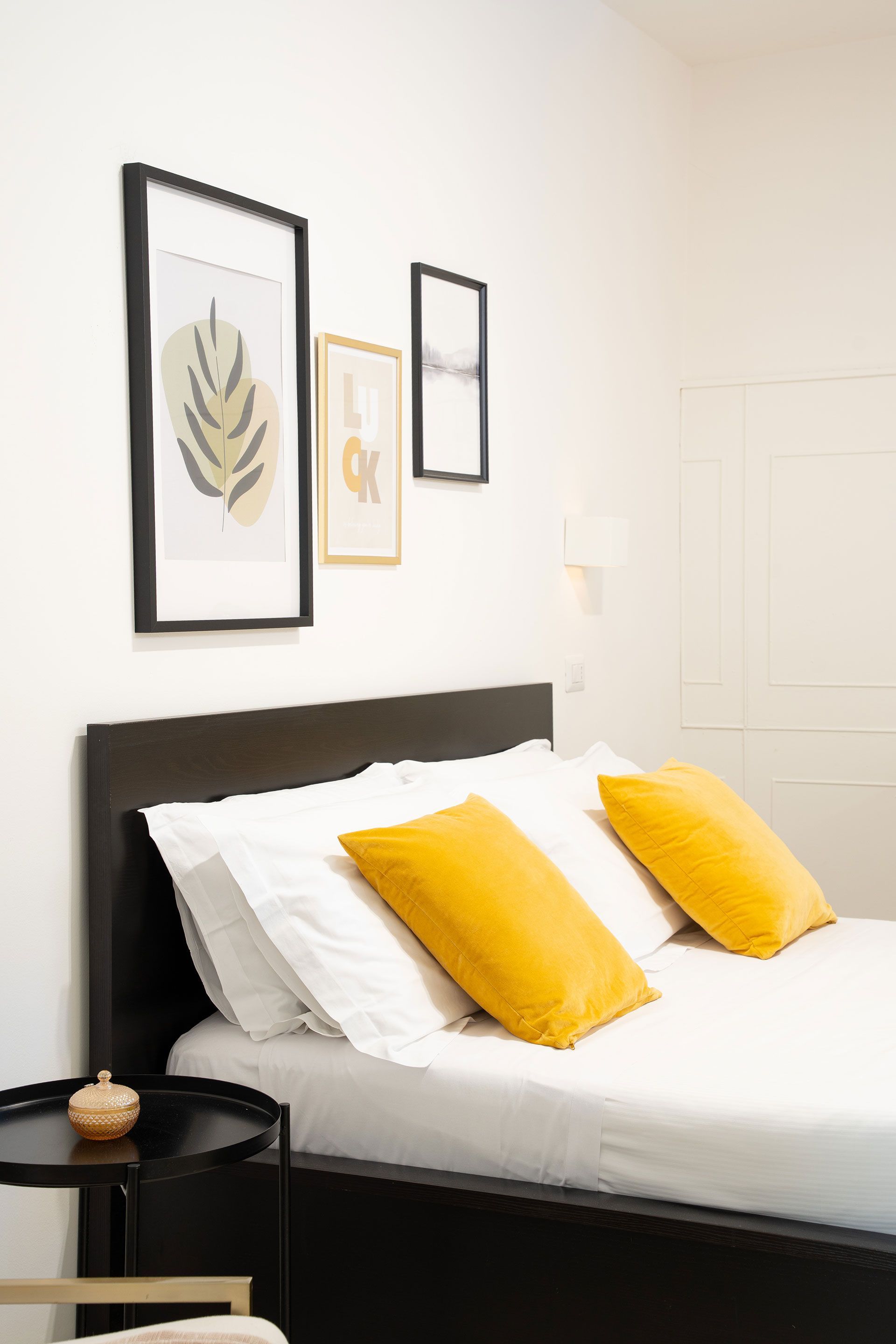 Camera da letto minimalista con lenzuola bianche, struttura del letto nera e cuscini decorativi gialli.