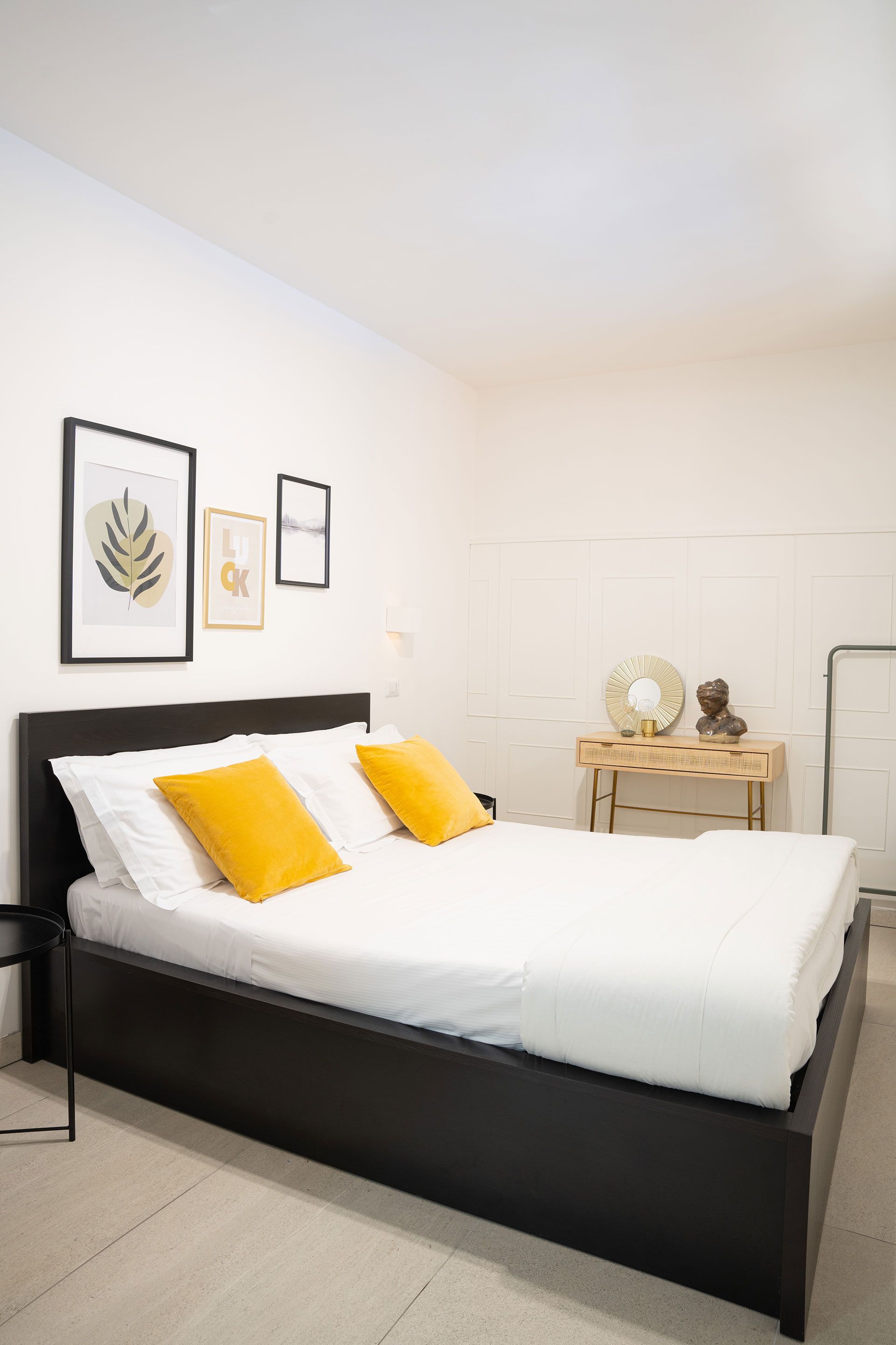 Camera da letto luminosa con letto nero, lenzuola bianche, cuscini gialli e quadri appesi alla parete.