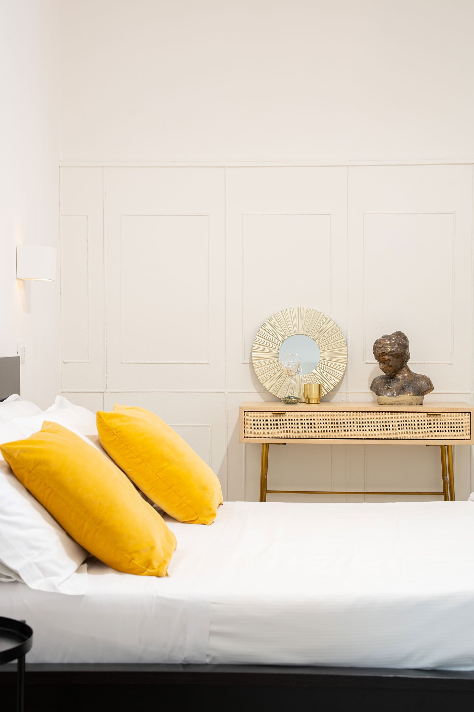 Camera da letto luminosa con letto bianco, cuscini gialli e una piccola consolle con specchio e busto scolpito.