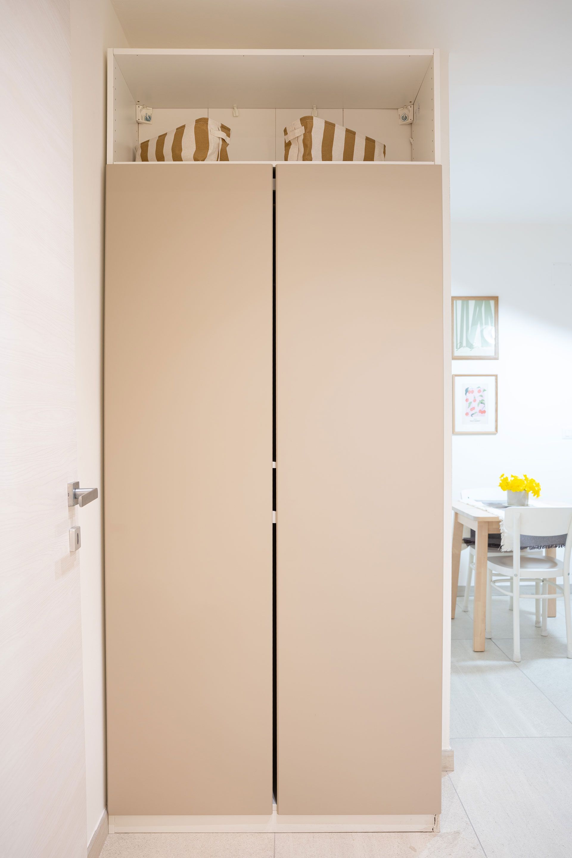 Armadio alto beige in una porta, con ripiani decorativi visibili sopra e zona pranzo sullo sfondo.