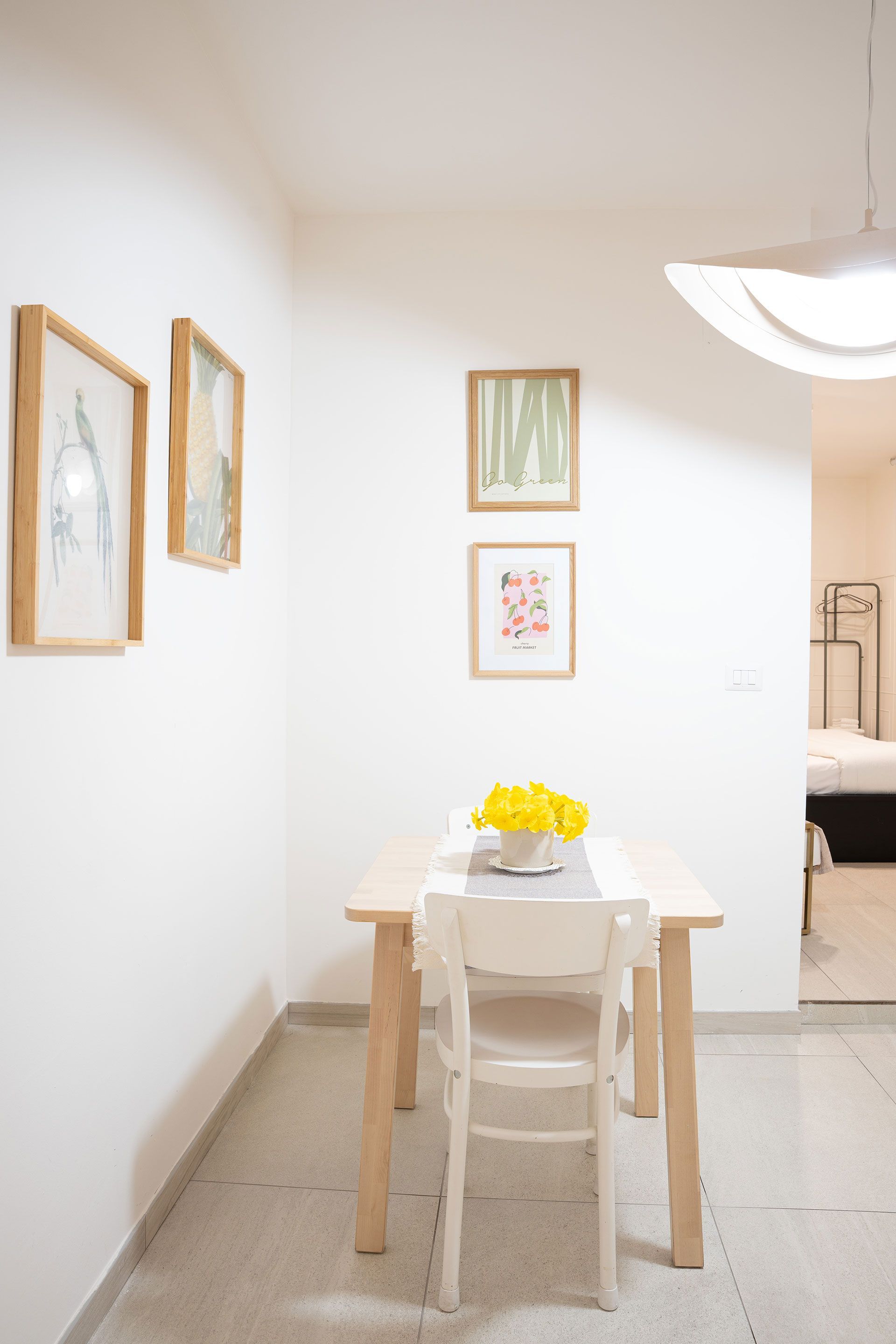 Luminosa zona pranzo con un tavolino bianco, sedie, quadri incorniciati e un vaso di fiori gialli in uno stretto corridoio.