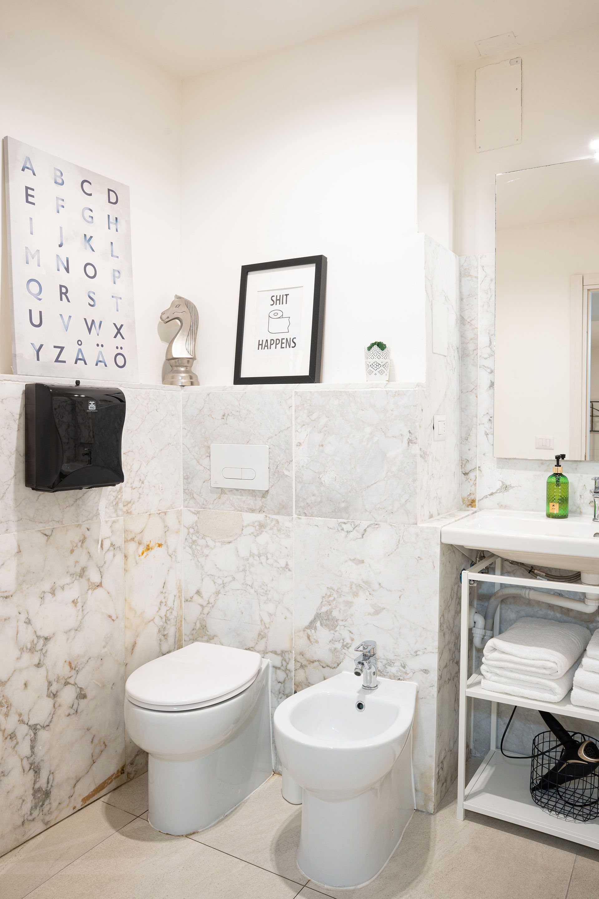 Piccolo bagno bianco con WC, bidet, pareti in marmo, un quadro con la tabella optometrica e un lavabo con mensole.