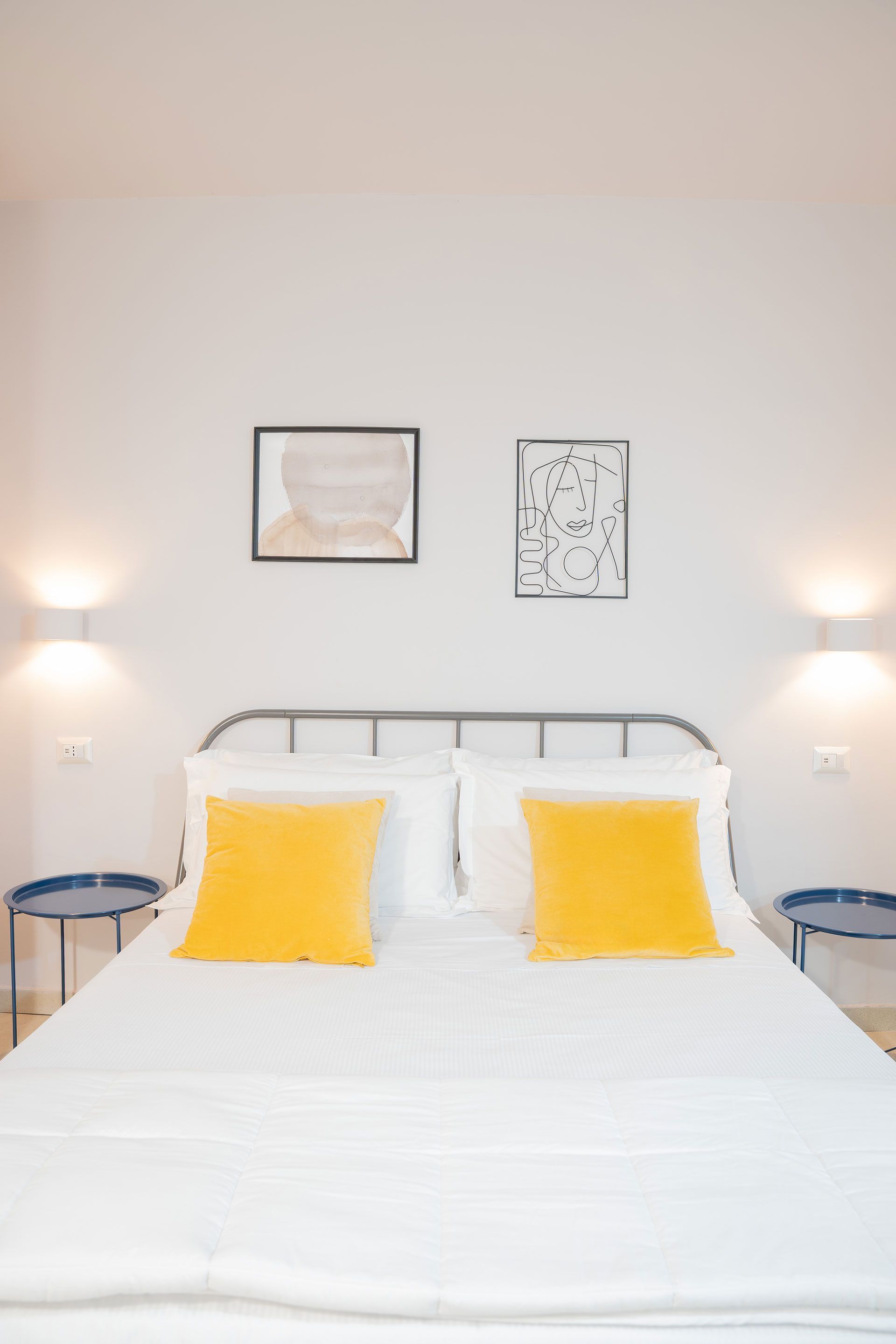 Camera da letto minimalista con lenzuola bianche, cuscini gialli, quadri alle pareti e lampade da comodino.