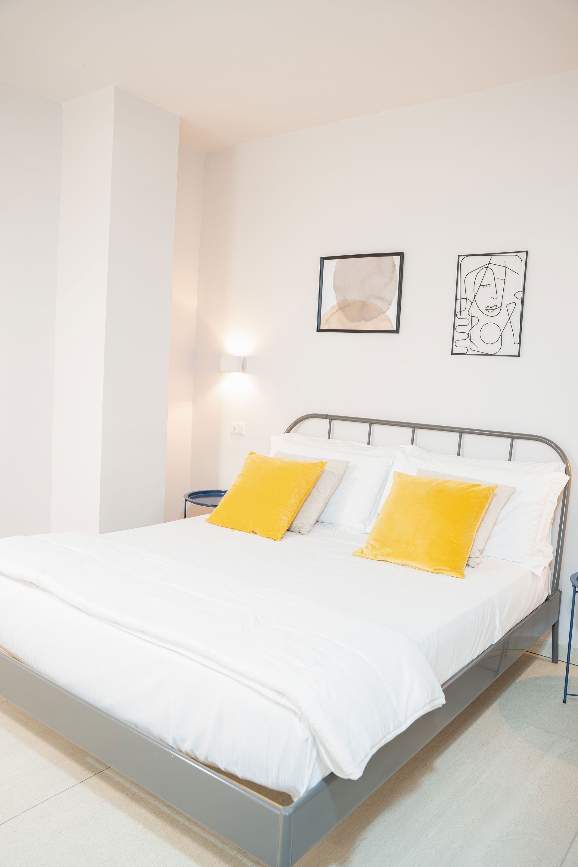 Camera da letto luminosa e bianca con letto, cuscini gialli e semplici quadri alle pareti.