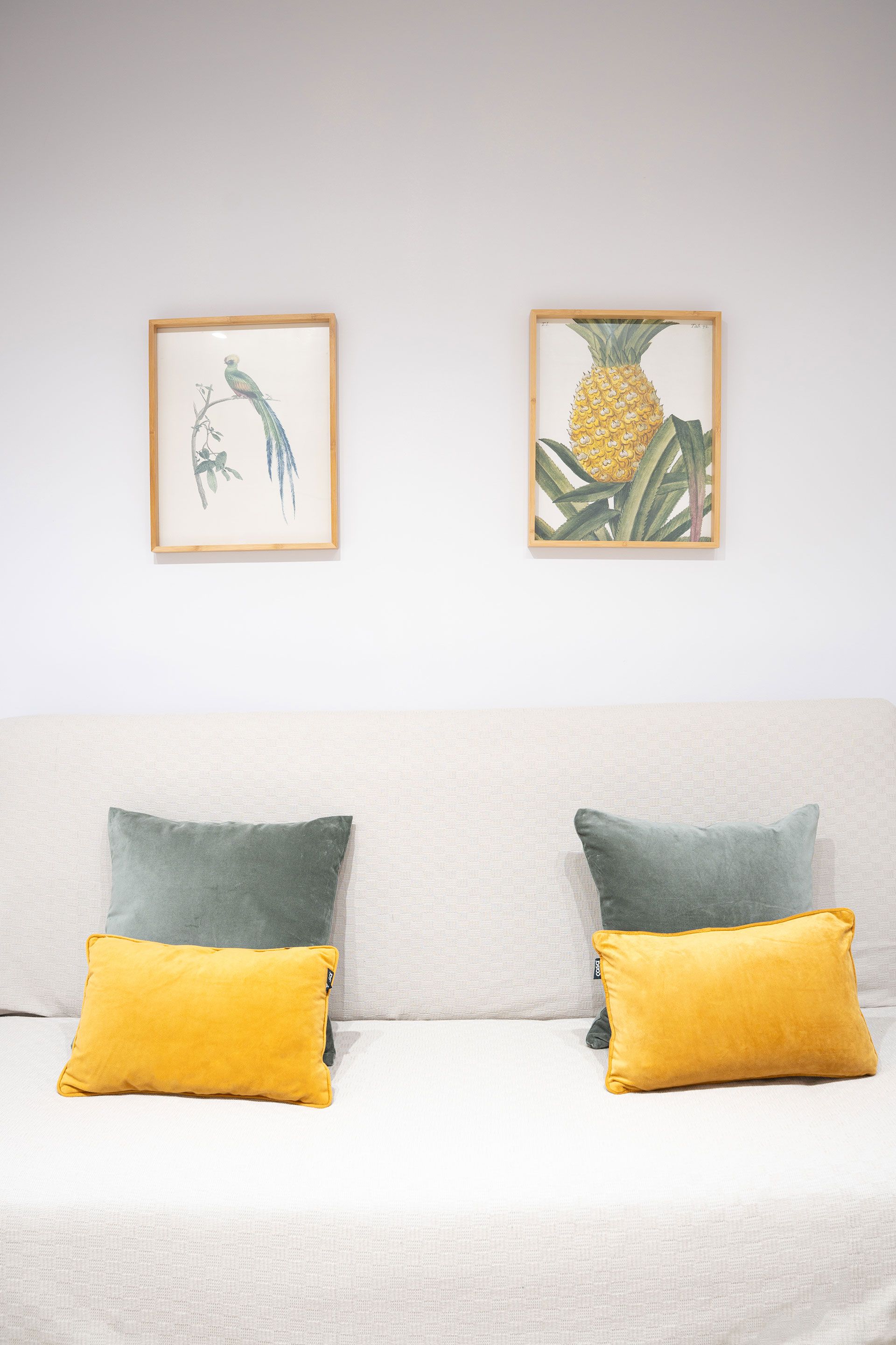 Soggiorno minimalista con divano bianco, cuscini verdi e gialli e due stampe botaniche incorniciate sopra.