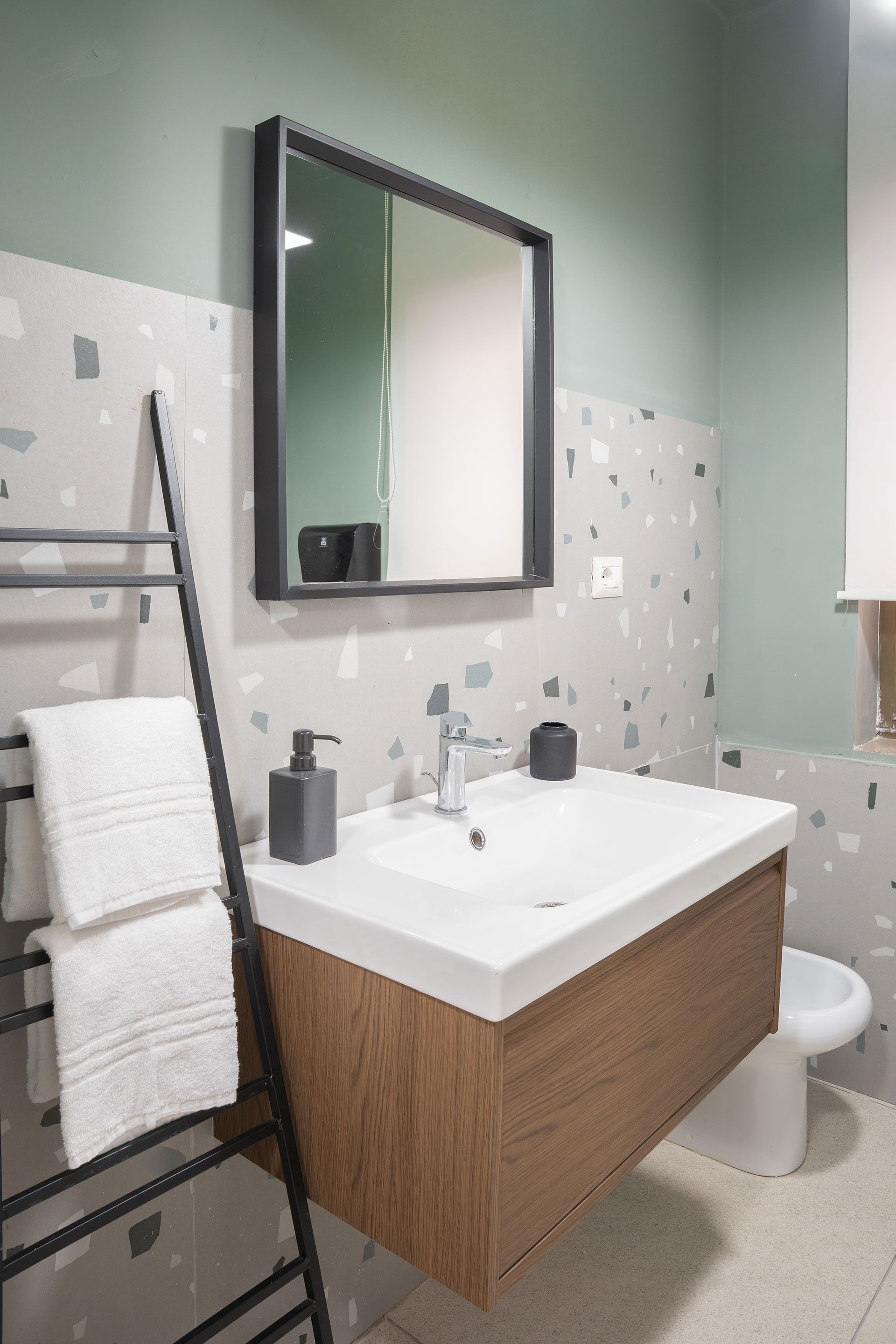 Mobile bagno moderno con lavabo bianco, mobiletto in legno, specchio, scaletta portasciugamani e pareti verdi.