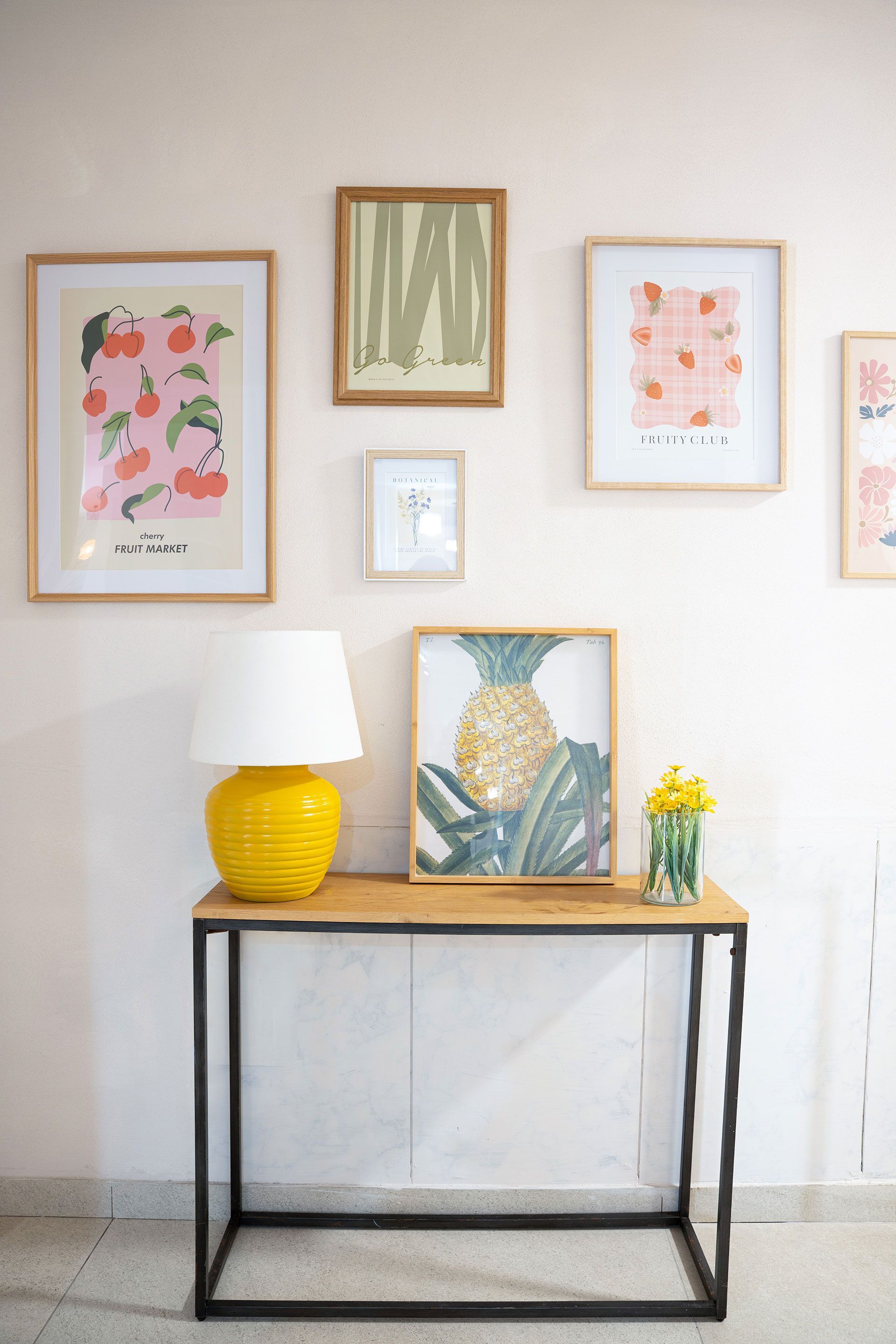 Stampe botaniche incorniciate sopra una consolle con lampada, vaso e fiori in una stanza luminosa.