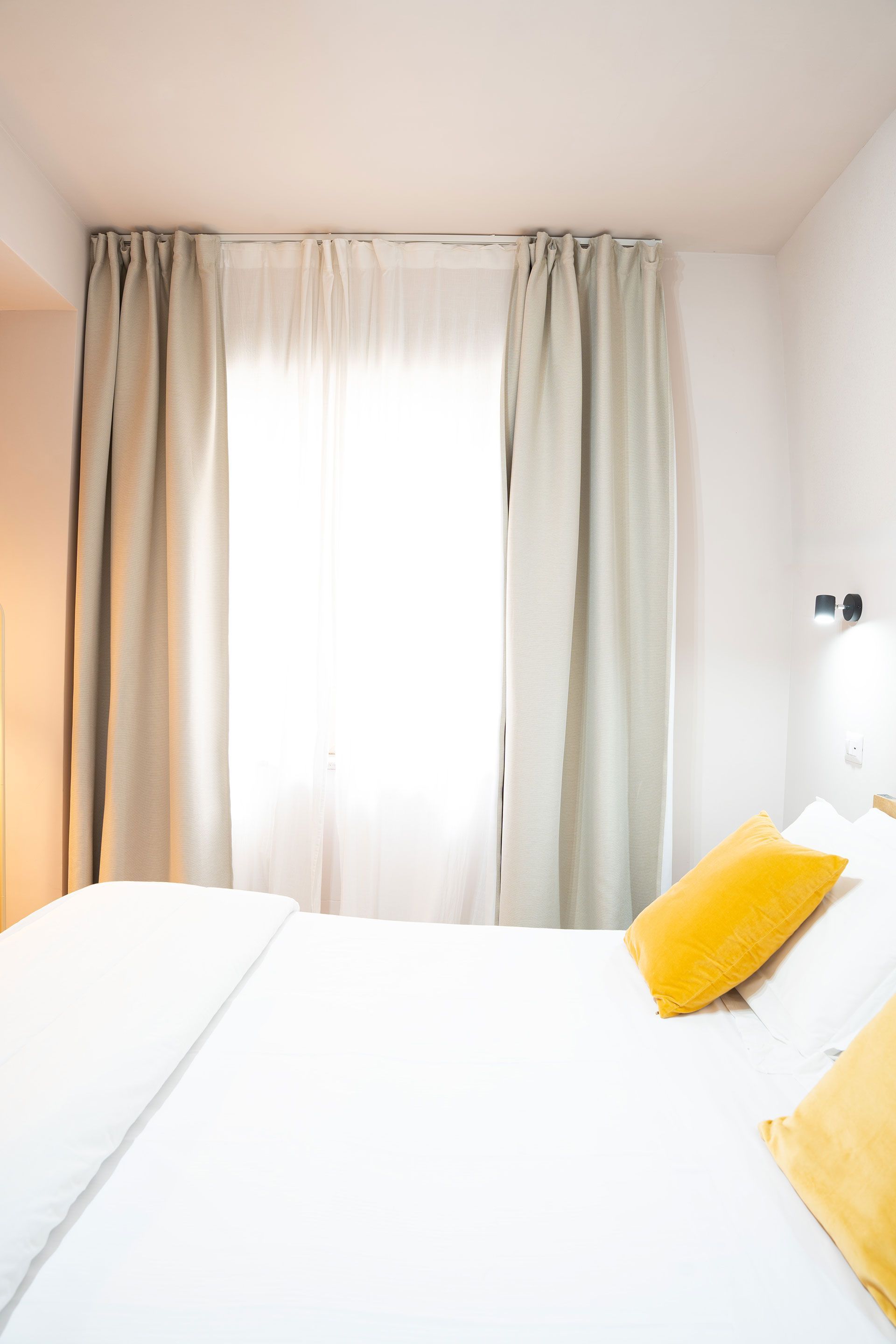 Camera da letto luminosa con lenzuola bianche, cuscini gialli e tende beige lunghe fino al pavimento su una finestra