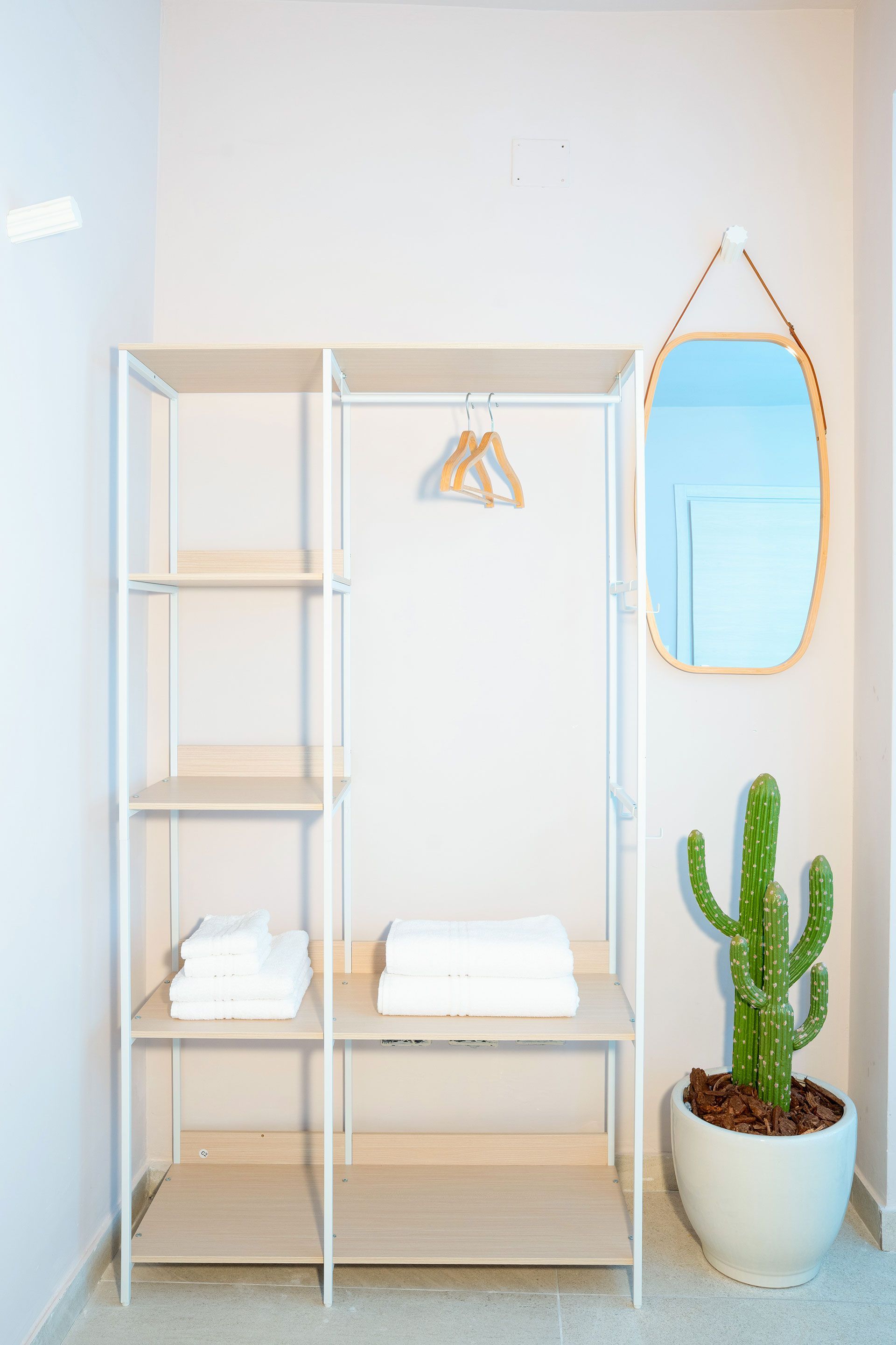 Armadio minimalista bianco con ripiano, appendiabiti, specchio, asciugamani piegati e cactus in vaso.
