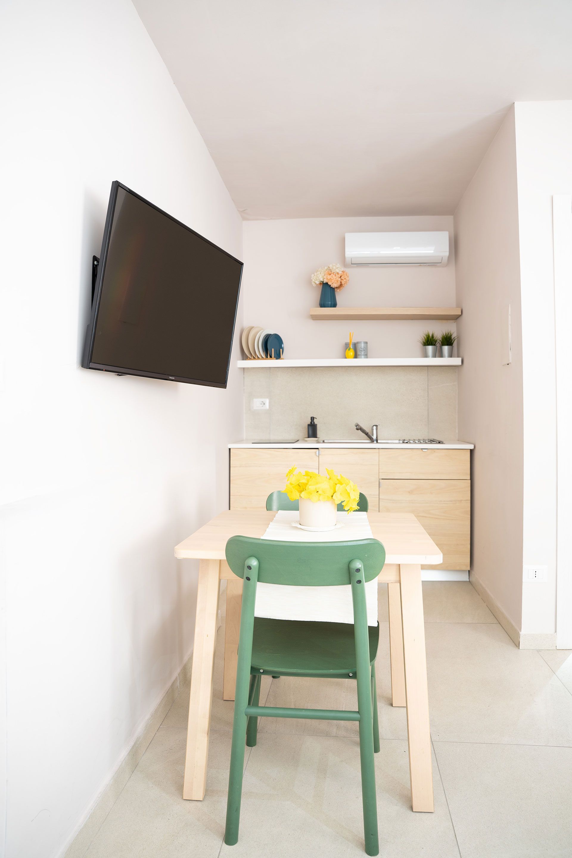 Piccolo angolo pranzo con TV a parete, tavolo in legno chiaro, sedia verde e mensole sospese su pareti beige.