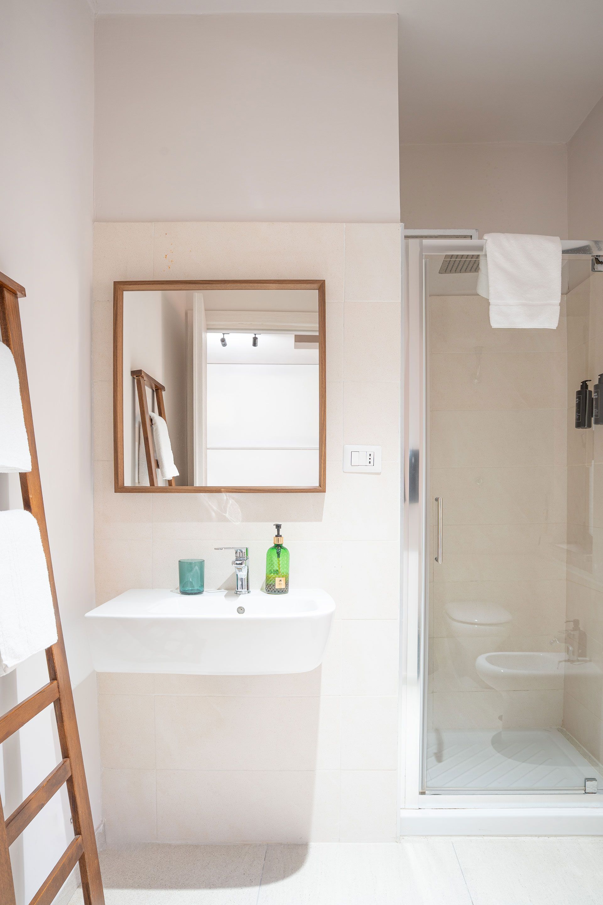 Piccolo bagno con lavandino, specchio, doccia in vetro e pareti bianche; i prodotti da bagno sono appoggiati sul lavandino.