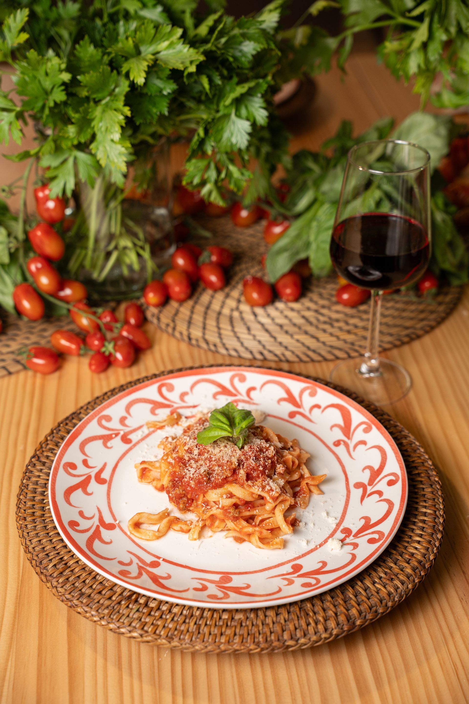 Pasta in una ciotola rossa e bianca su una tovaglietta intrecciata, con un calice di vino e pomodorini su un tavolo rustico.