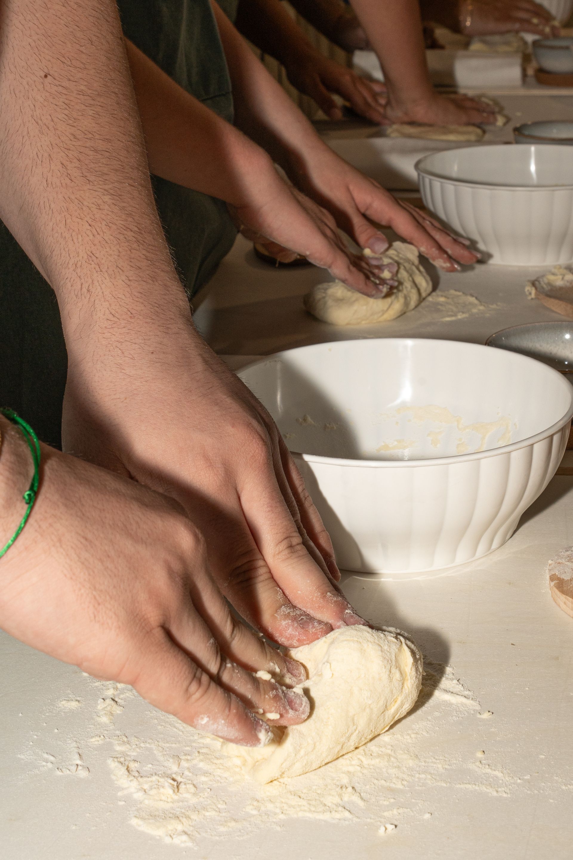 Le mani impastano la pasta su un piano di lavoro infarinato, accanto a delle ciotole bianche.