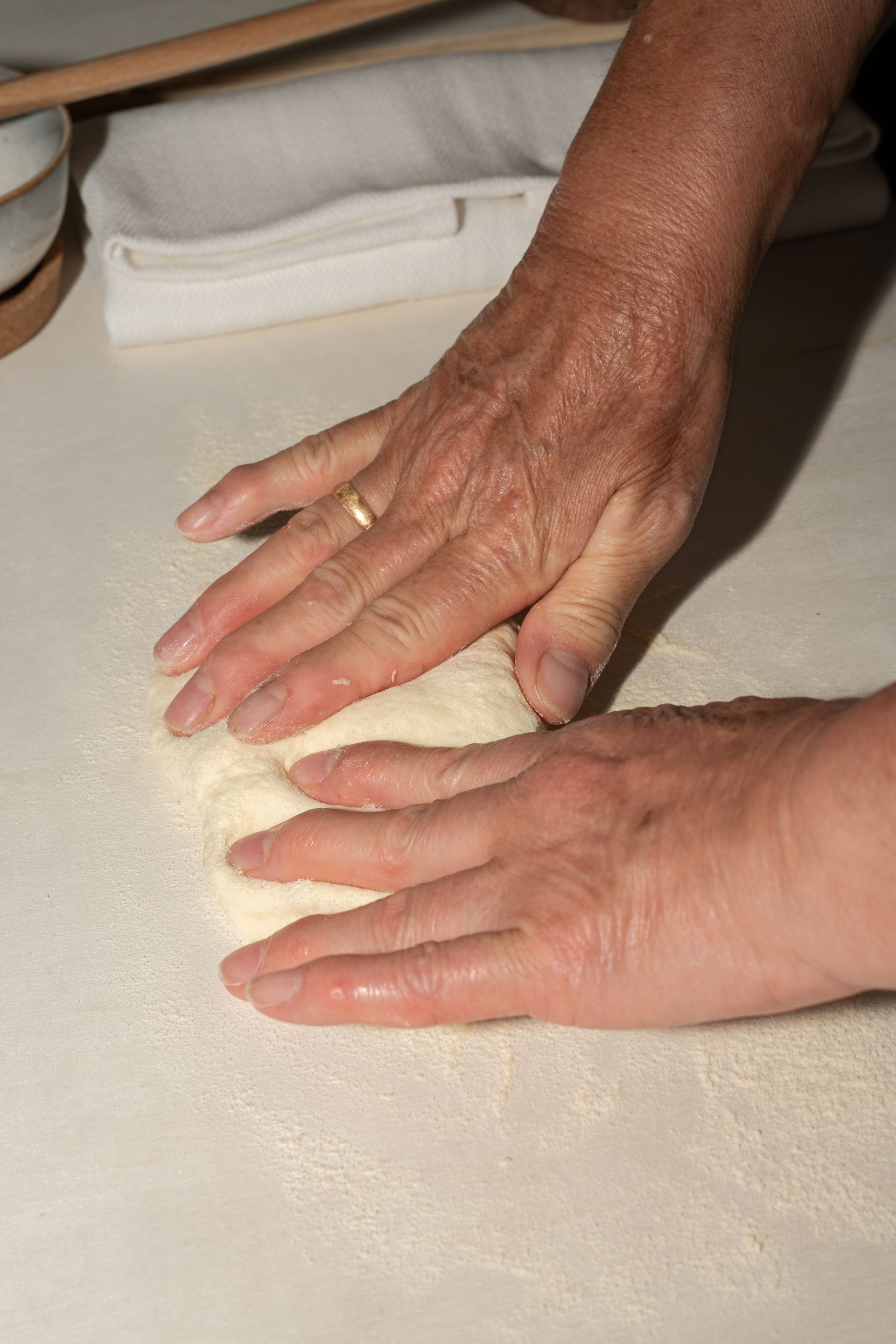 Mani che impastano la pasta su un piano di lavoro infarinato, con un panno sullo sfondo.
