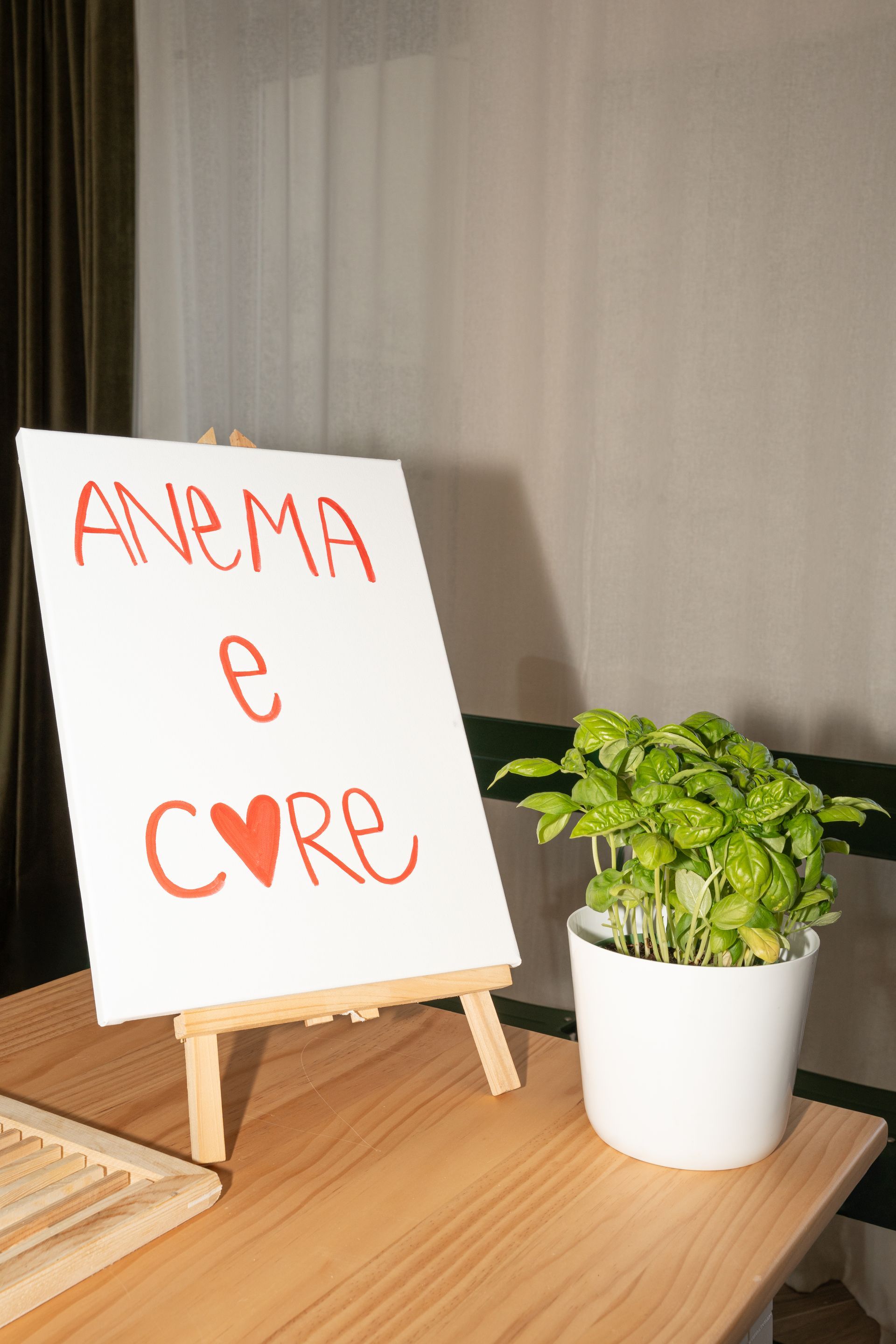 Un cartello scritto a mano con la scritta “ANEMA E CORE” su un piccolo cavalletto accanto a una pianta in vaso su un tavolo di legno.