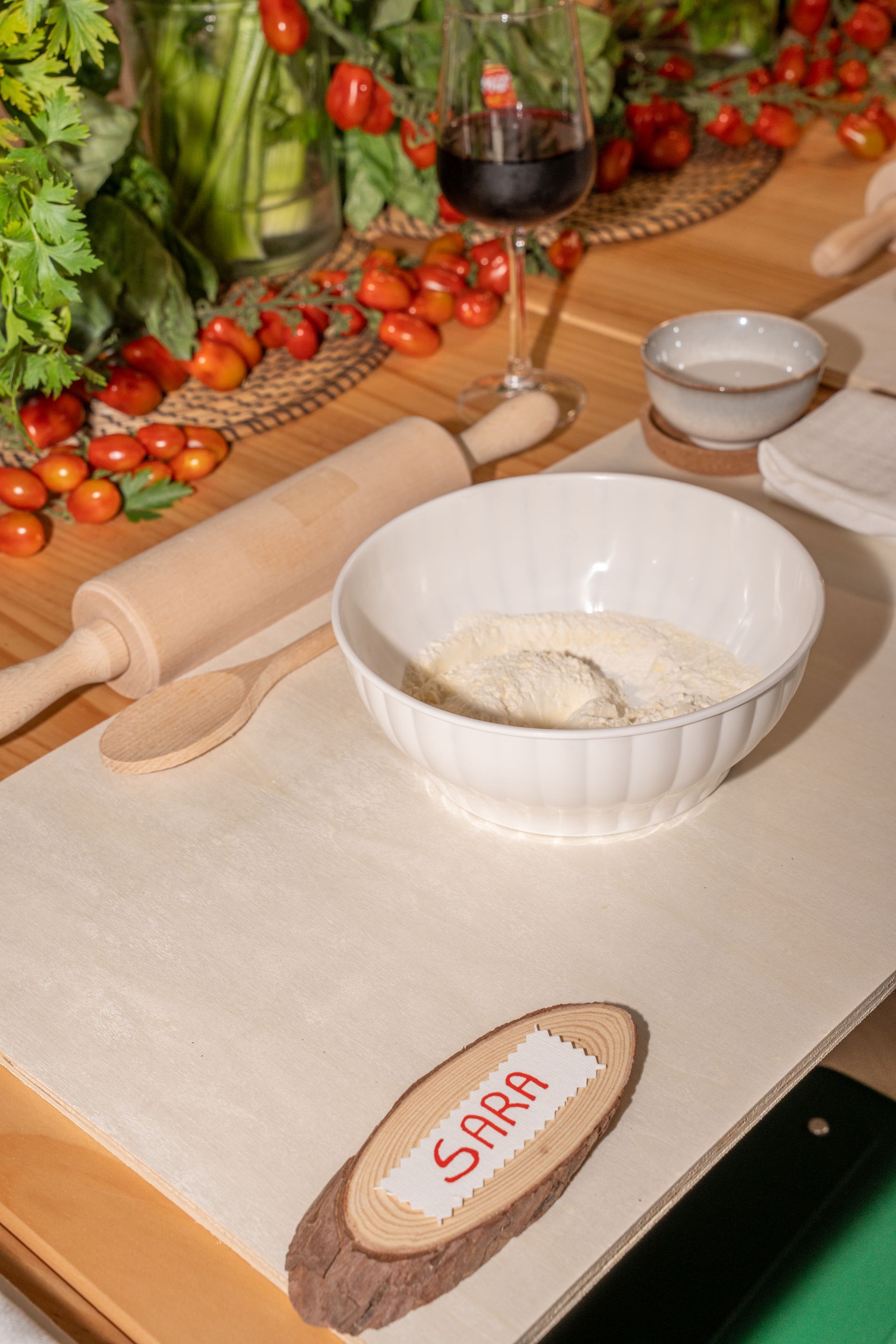 Lievito madre su un piano di lavoro infarinato, con pomodori, mattarello e bicchiere di vino sullo sfondo.
