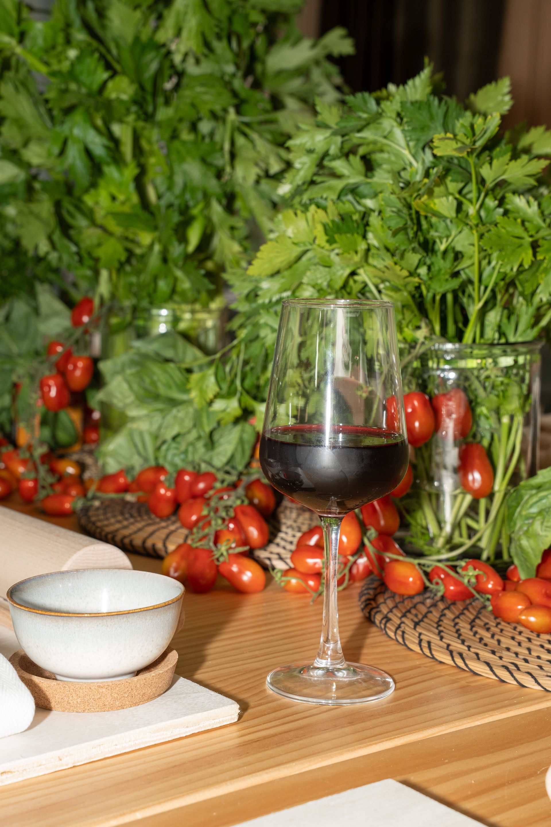 Un calice di vino tra erbe aromatiche fresche, pomodorini e ciotole su un tavolo di legno.