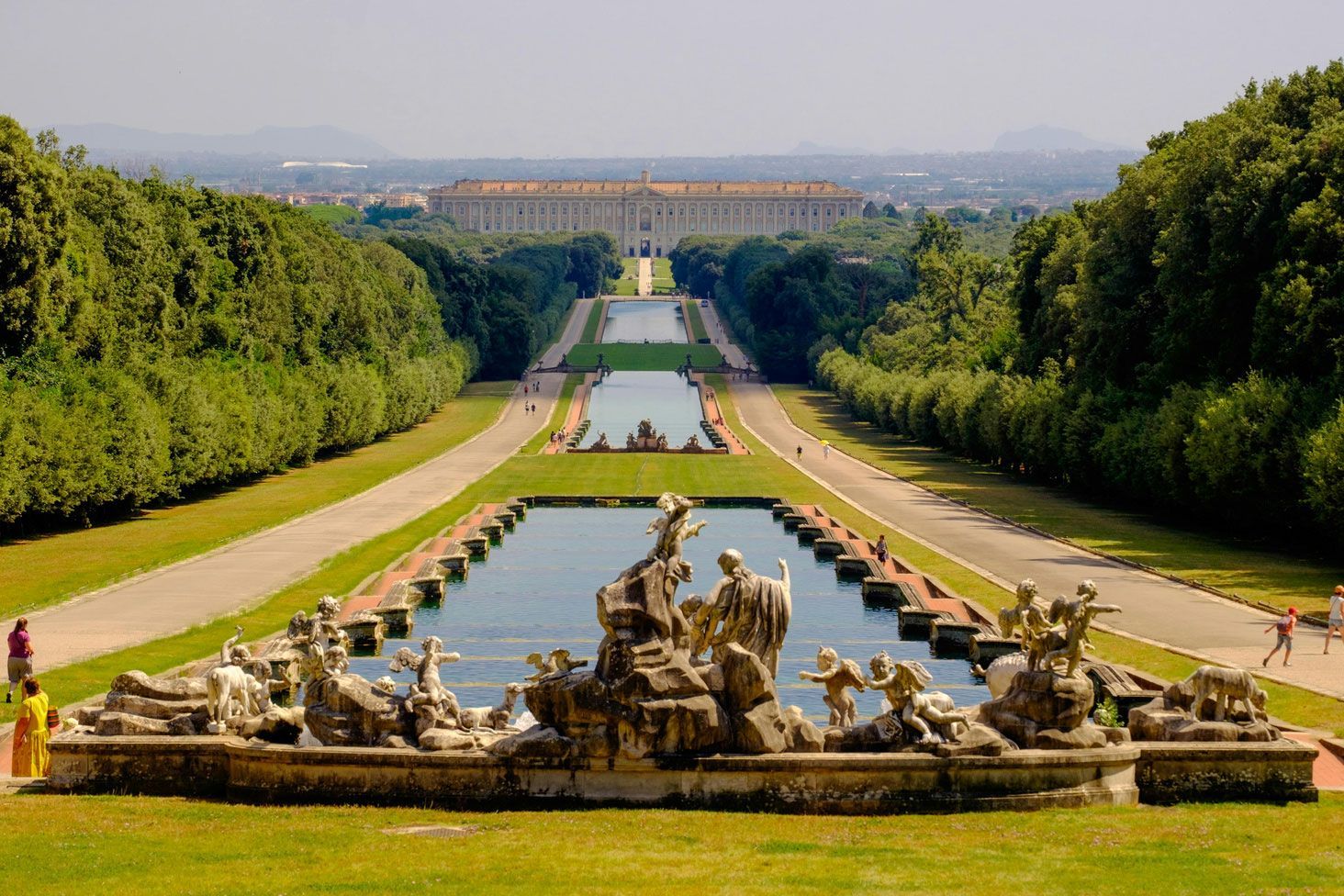 Giardino formale con lunga vasca riflettente, sculture di fontane ornate e alberi che costeggiano una vista maestosa sul palazzo.